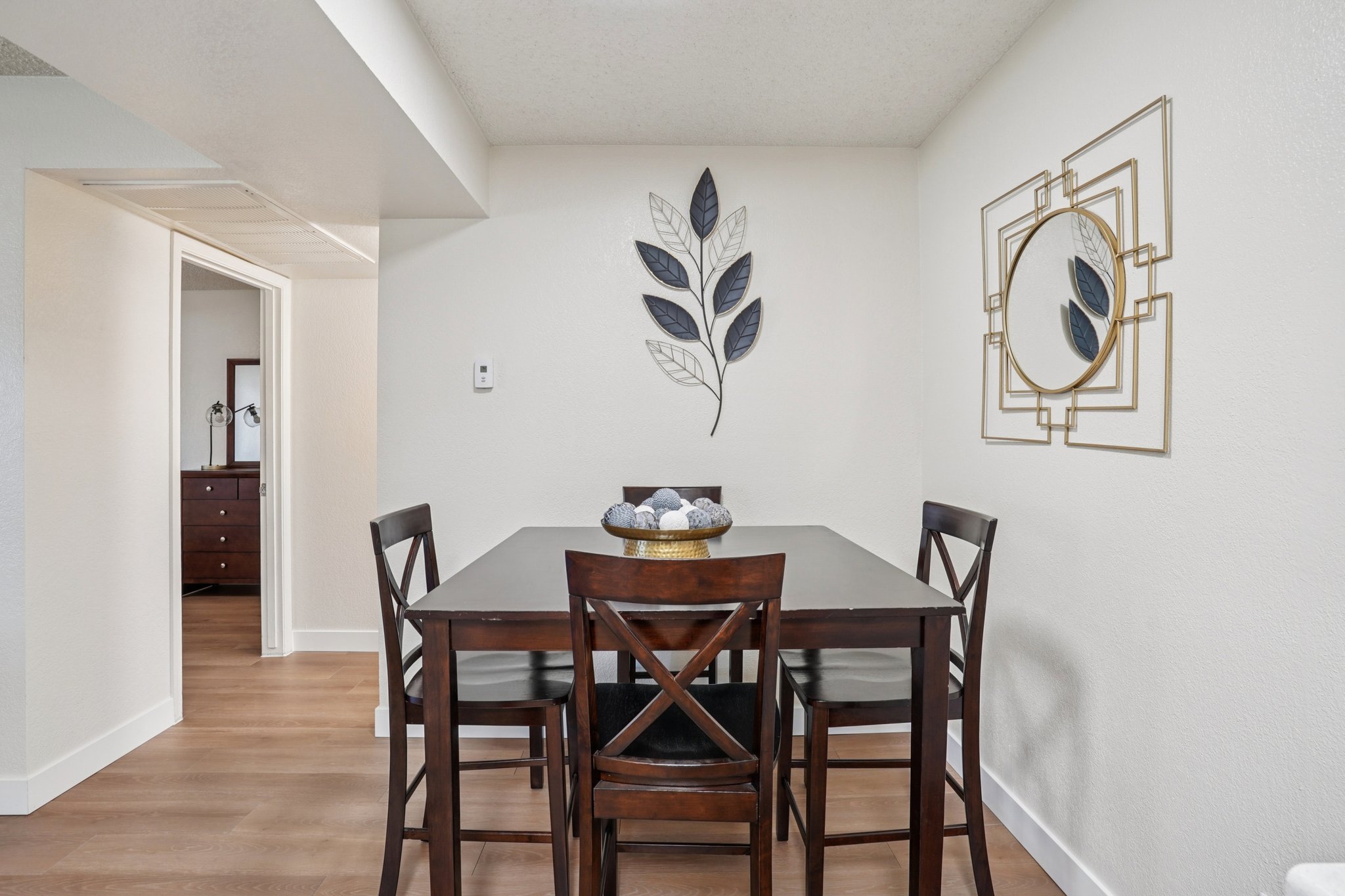 A small dining area featuring a dark wood table with four matching chairs. A decorative bowl with objects sits at the center of the table. The wall is adorned with a leaf-themed artwork and a round mirror. An open doorway leads to another room in the background, with light-colored walls and hardwood flooring.