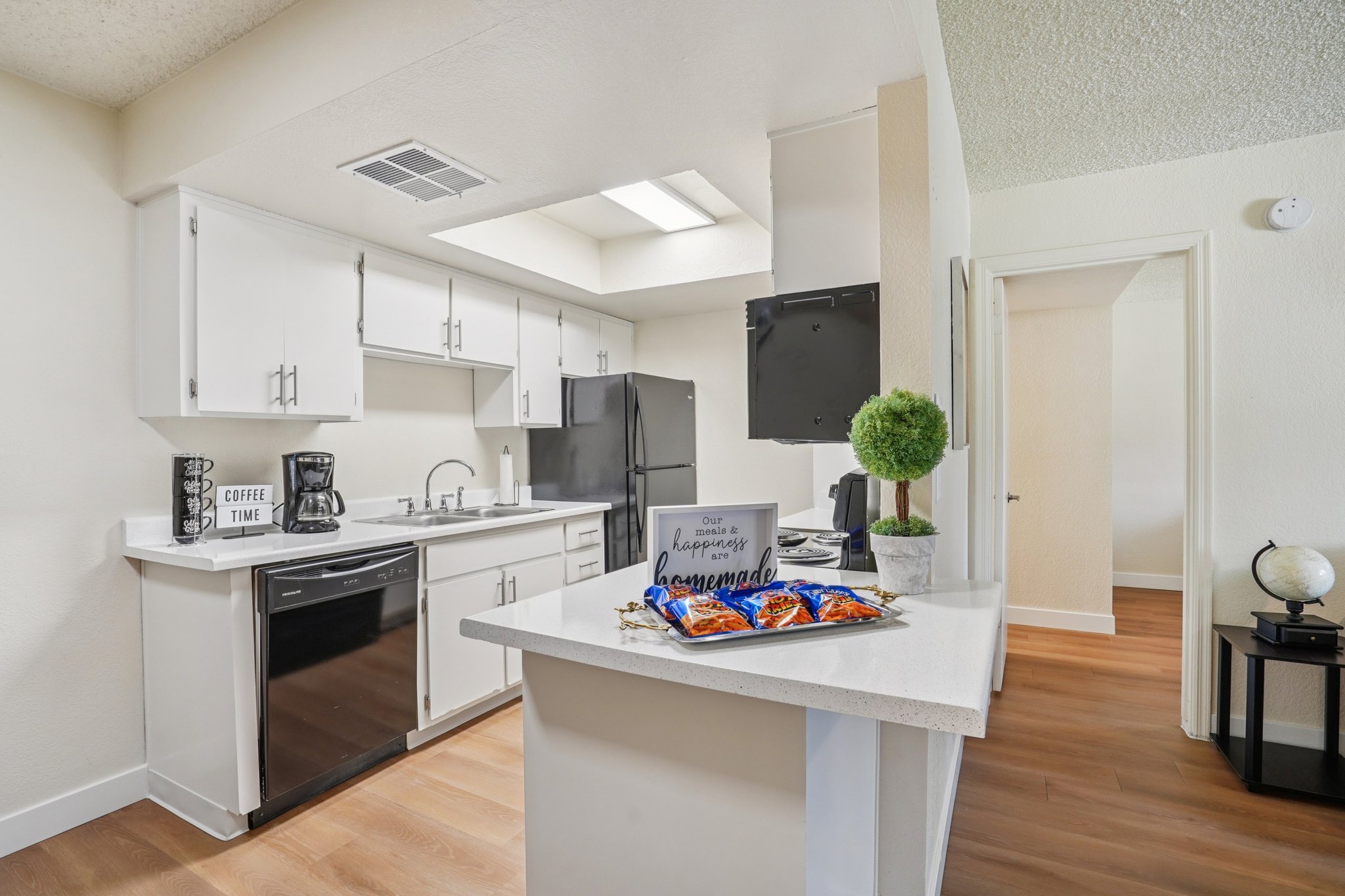 Modern kitchen featuring white cabinets, stainless steel appliances, and a black refrigerator. A small island with a quartz countertop displays a sign reading "Do what makes you happy" alongside a colorful snack platter. Natural light fills the space through a skylight, with wood-like flooring and neutral wall colors.