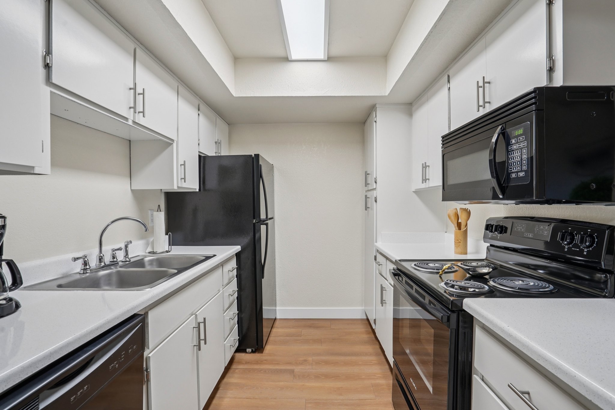 A modern kitchen featuring white cabinets, a black refrigerator, a black microwave, and an electric stove. The countertops are light-colored, and there is a double sink with a faucet. Wooden flooring complements the neutral wall color. A skylight provides natural light in the space.
