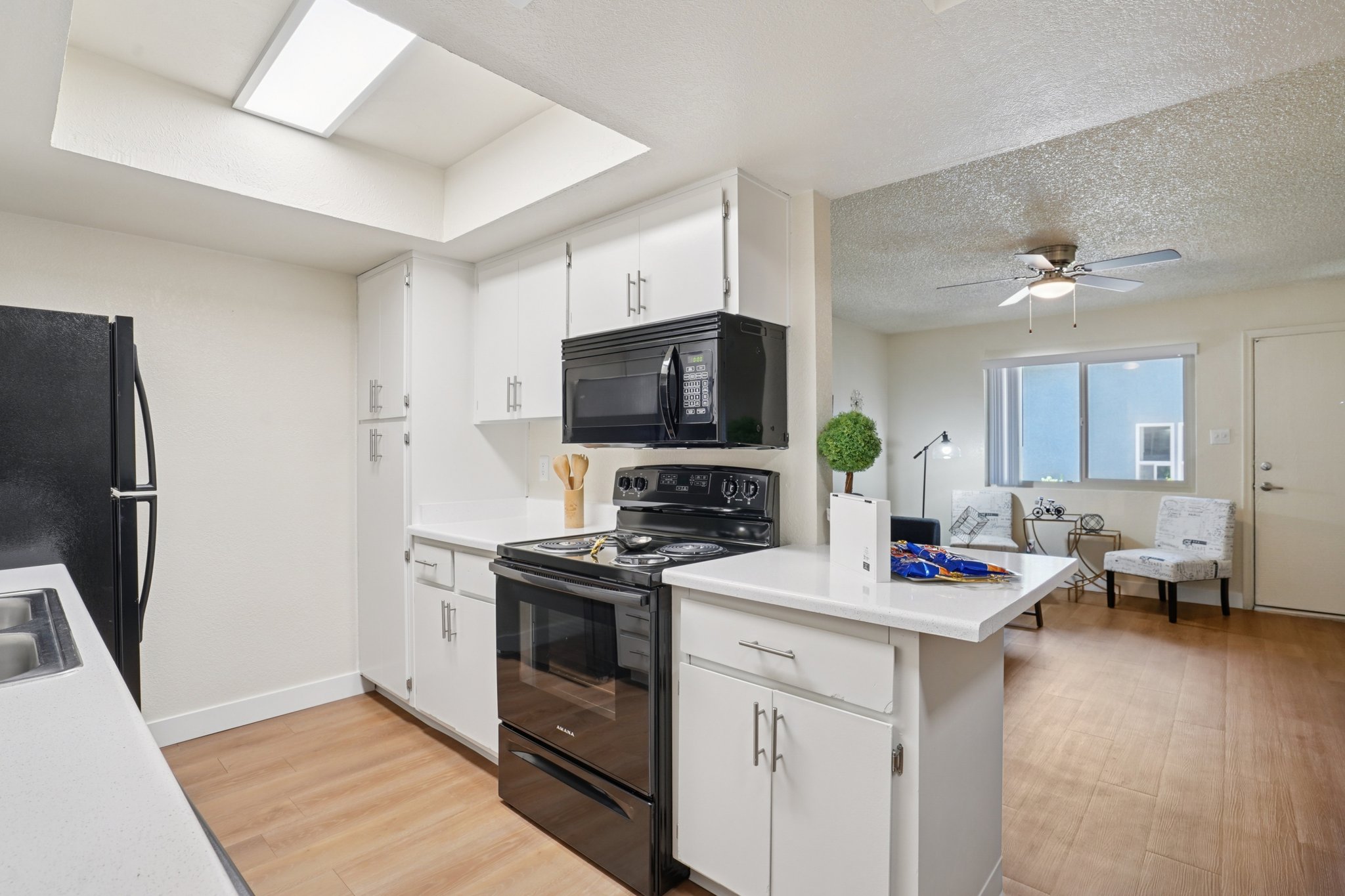 A modern kitchen featuring white cabinets, a black refrigerator, and a black stove with an overhead microwave. The countertops are light-colored with utensils and a small plant for decoration. In the background, a living area is visible with a ceiling fan and two chairs. Natural light comes through a nearby window.