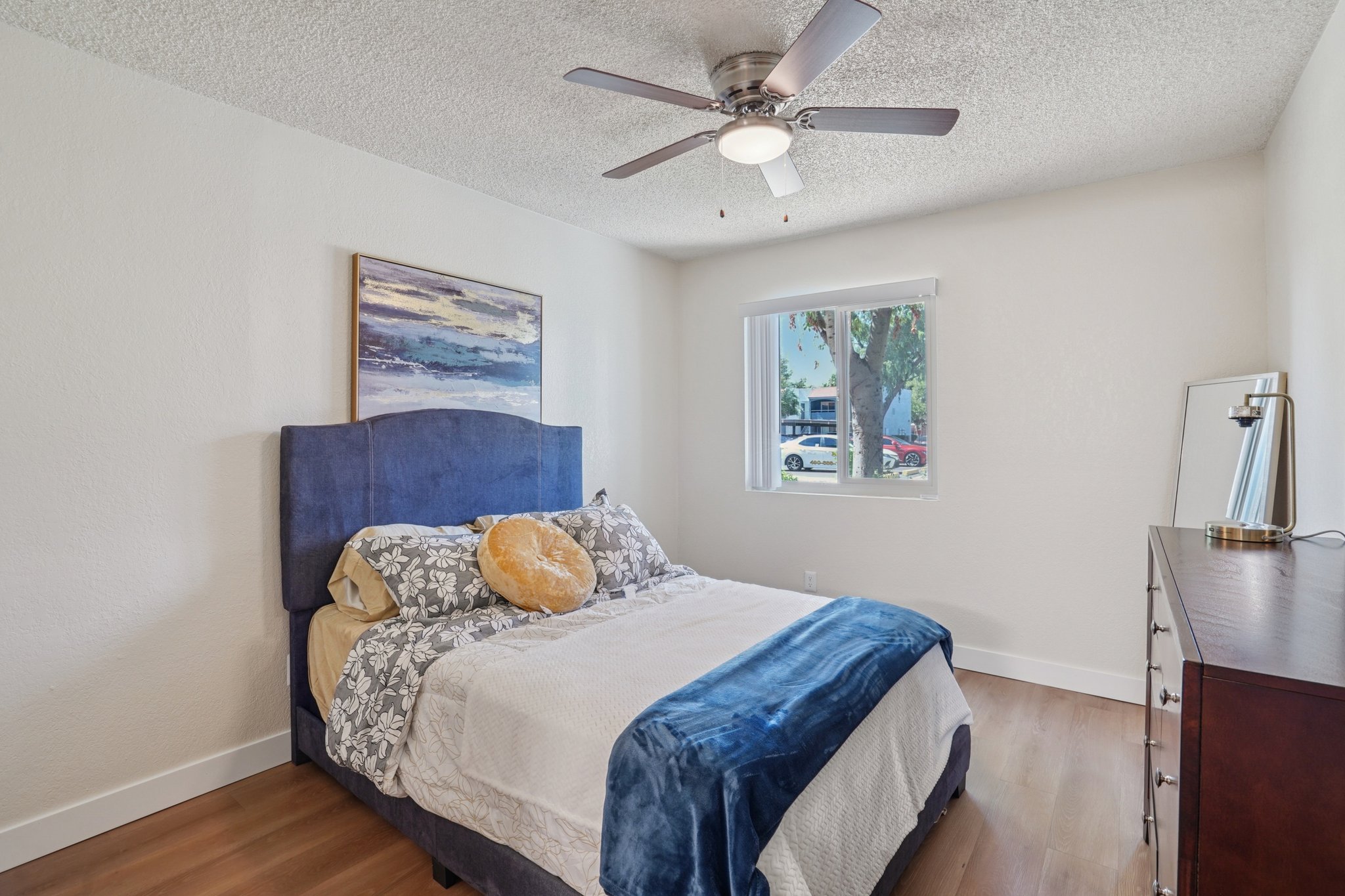 A cozy bedroom featuring a blue upholstered headboard with a patterned bedspread and a large round pillow. There is a ceiling fan, a painting on the wall, a window with blinds, and a wooden dresser. Soft natural light fills the room, creating a warm and inviting atmosphere.