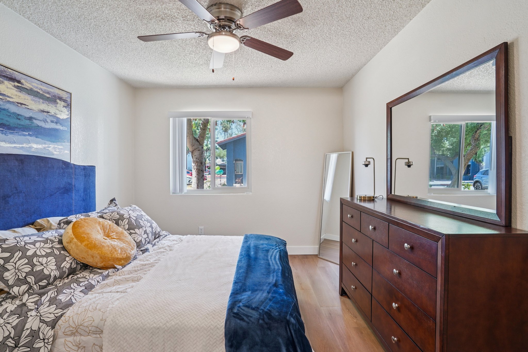 Cozy bedroom featuring a king-size bed with decorative pillows, a wooden dresser, a mirror, and a ceiling fan. Natural light enters through a window, highlighting a blue and white wall art piece. A large decorative pillow and a textured blanket add warmth to the space.
