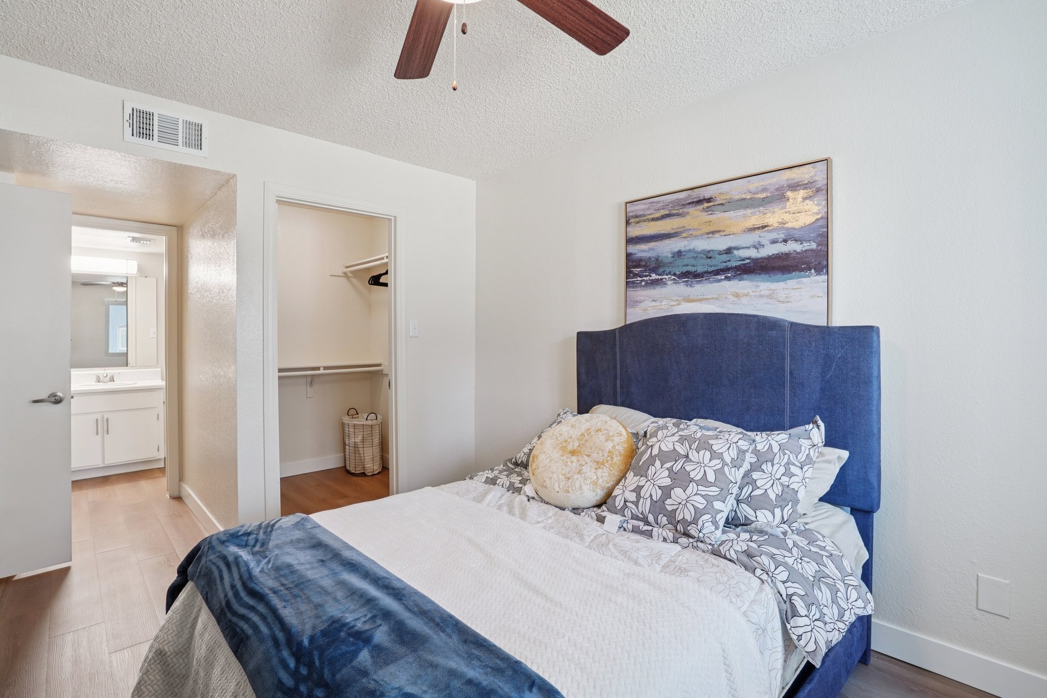A cozy bedroom featuring a blue upholstered bed with decorative pillows, a light-colored blanket, and a textured wall. There is a wooden fan on the ceiling, a closet space visible, and a bathroom area in the background. Natural light brightens the room, complemented by a modern abstract painting on the wall.