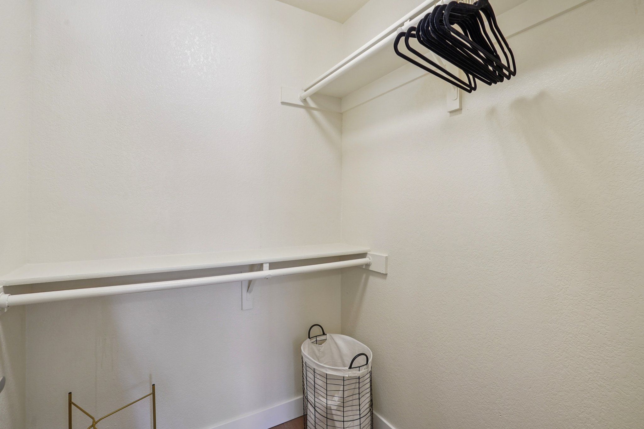 A neatly organized closet with a white wall and empty shelving. There are a few black hangers on the upper shelf and a wire laundry basket on the floor. The overall space is bright and clean, emphasizing minimalism and functionality.