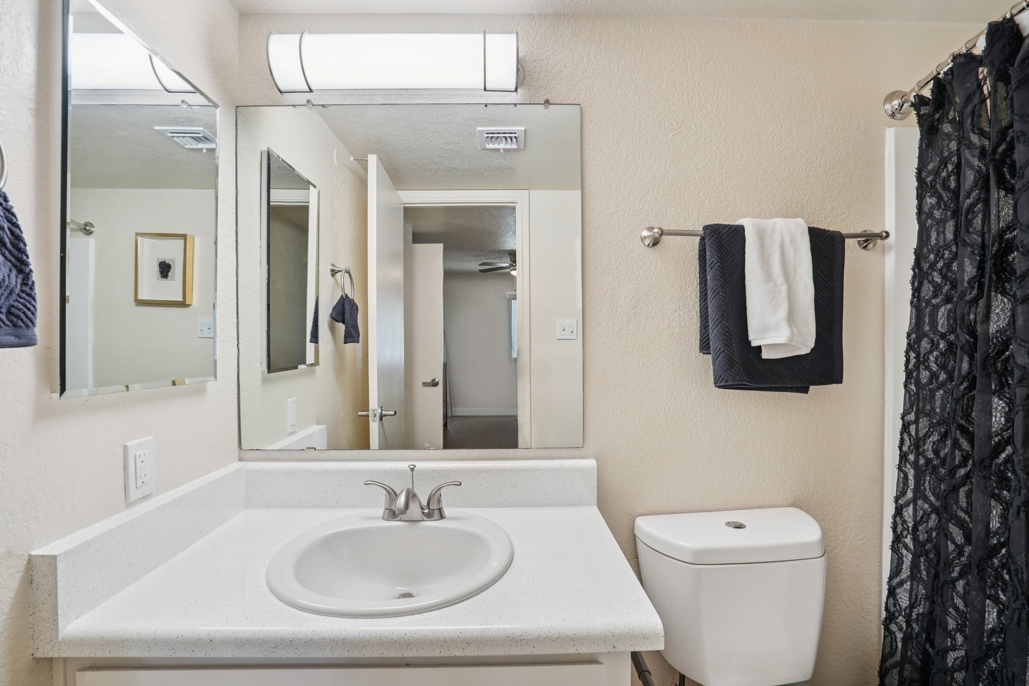 A clean, modern bathroom featuring a white sink with a polished faucet, a toilet, and a large mirror above the sink. The walls are light-colored, and there's a decorative black shower curtain. Towels are neatly hung on a towel rack, and an open door leads to another room in the background.