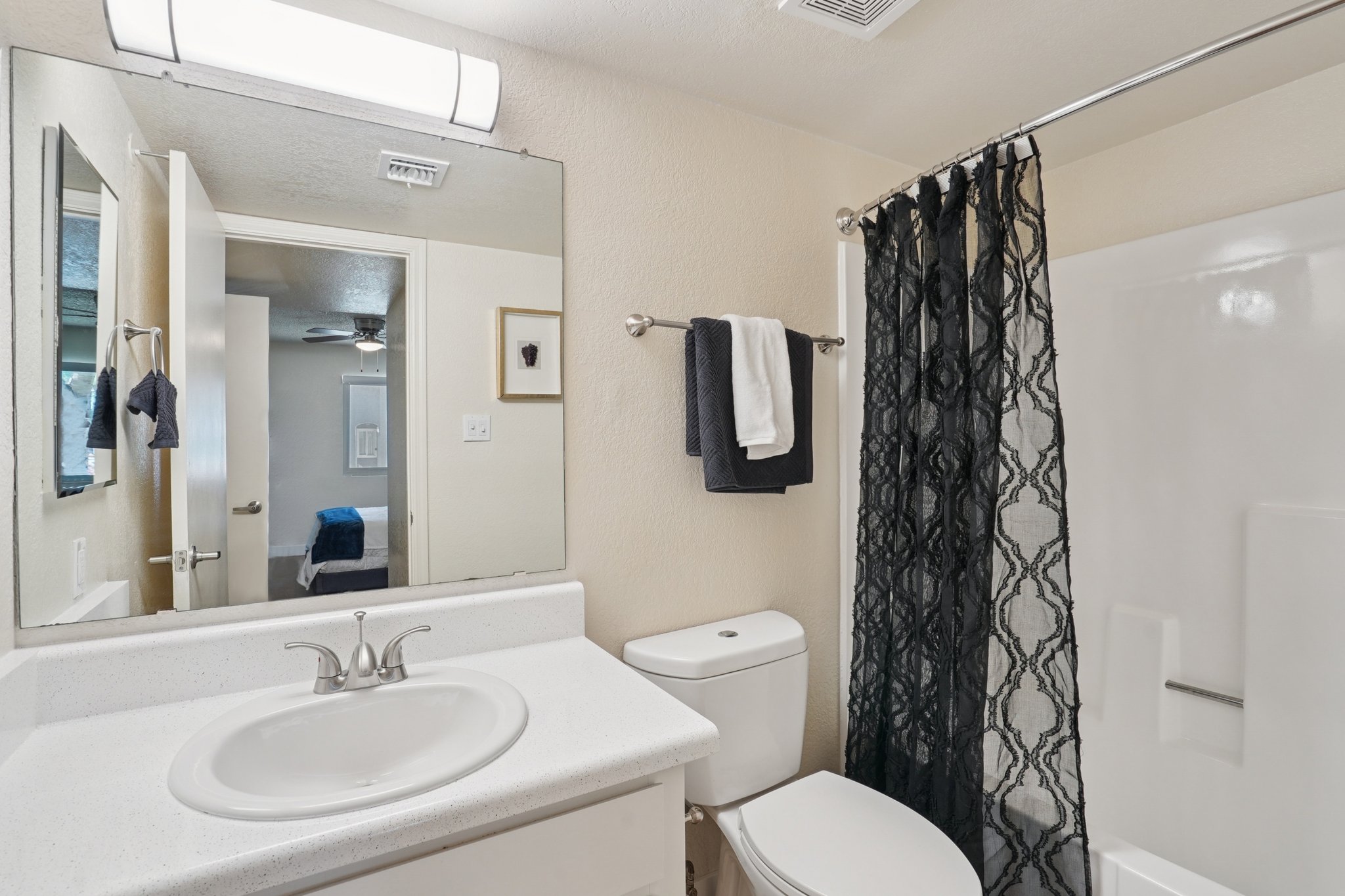 A modern bathroom featuring a white countertop sink, a toilet, and a shower with a black curtain. There’s a large mirror above the sink, and a towel rack with two towels. The walls are painted a light color, and a door is visible in the background, leading to another room.