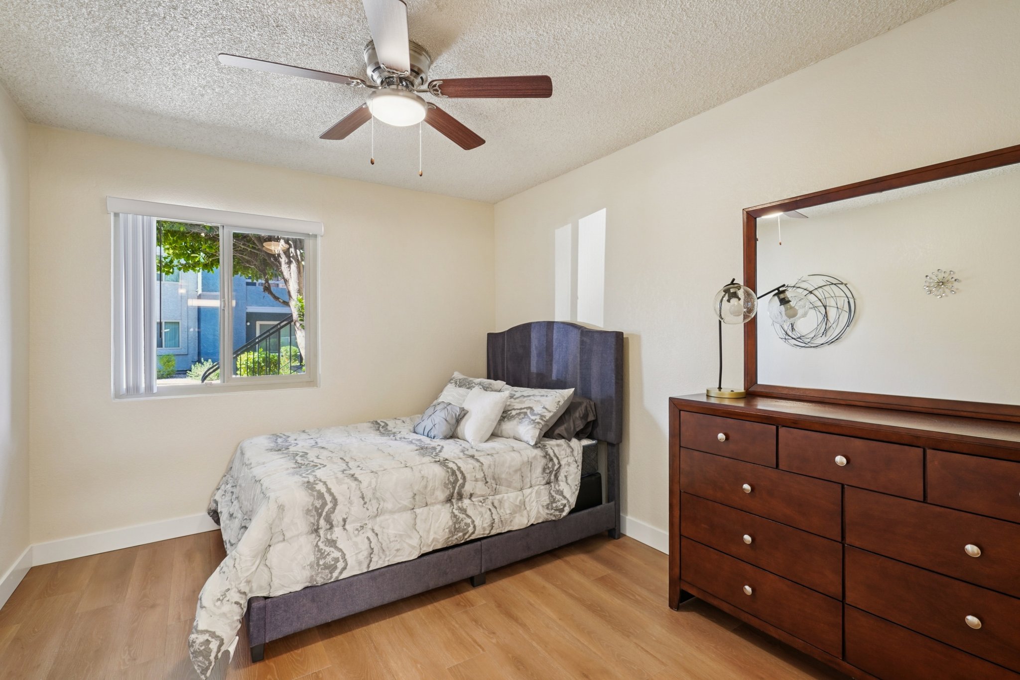 A cozy bedroom featuring a bed with a gray and white patterned comforter, a wooden dresser with multiple drawers, and a ceiling fan. A window allows natural light to enter, showcasing greenery outside. The walls are painted a soft beige, creating a warm atmosphere.