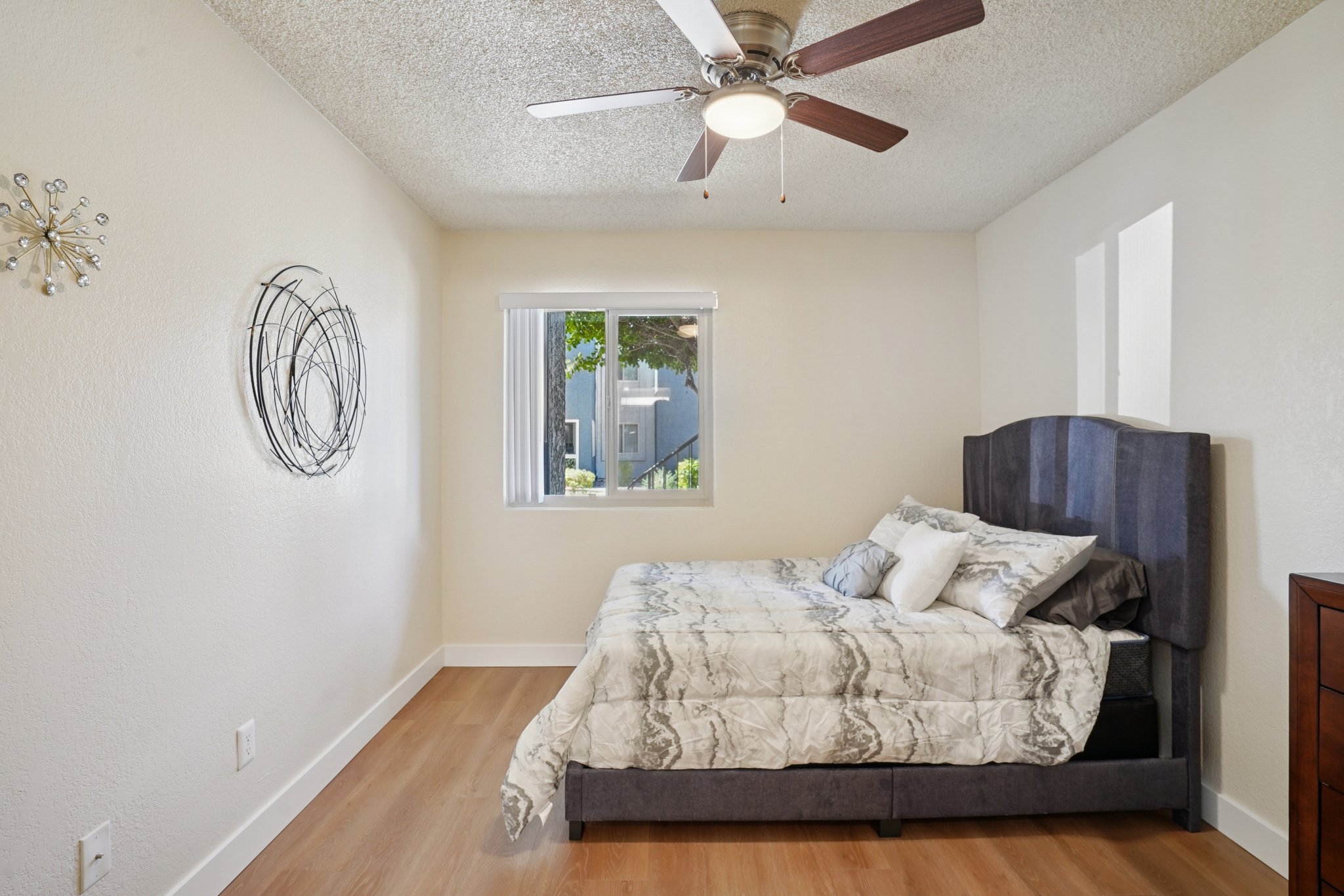 Cozy bedroom featuring a dark gray upholstered bed with a patterned comforter, a ceiling fan, and a window allowing natural light. The walls are light-colored, and there's a decorative wall hanging near the bed. The flooring is wooden, creating a warm and inviting atmosphere.