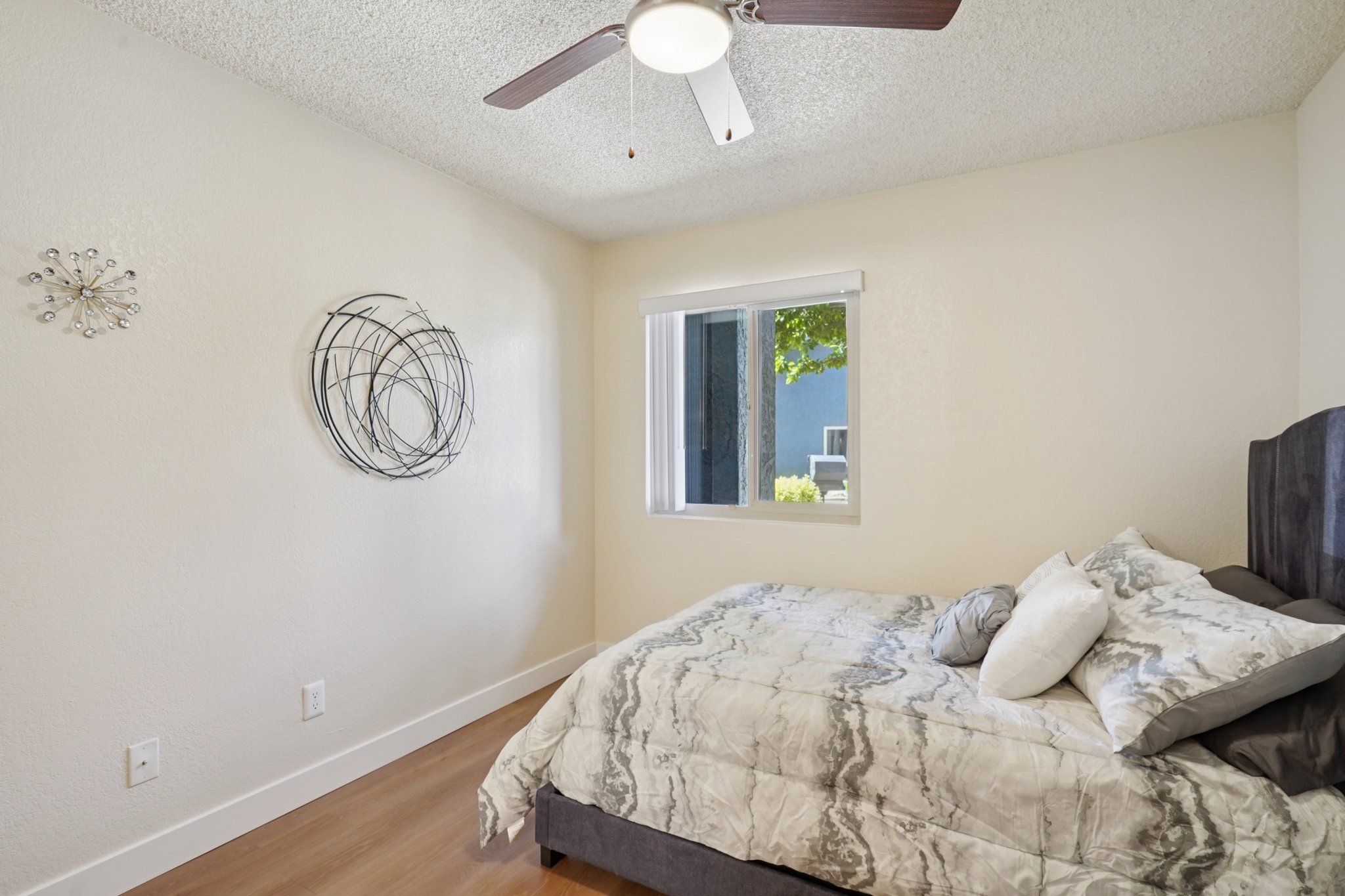 A cozy bedroom featuring a bed with a marble-patterned comforter and decorative pillows. The room has light-colored walls, a ceiling fan, and a window allowing natural light. A circular wall art piece adds visual interest to the space. The floor is made of light wood, creating a warm atmosphere.