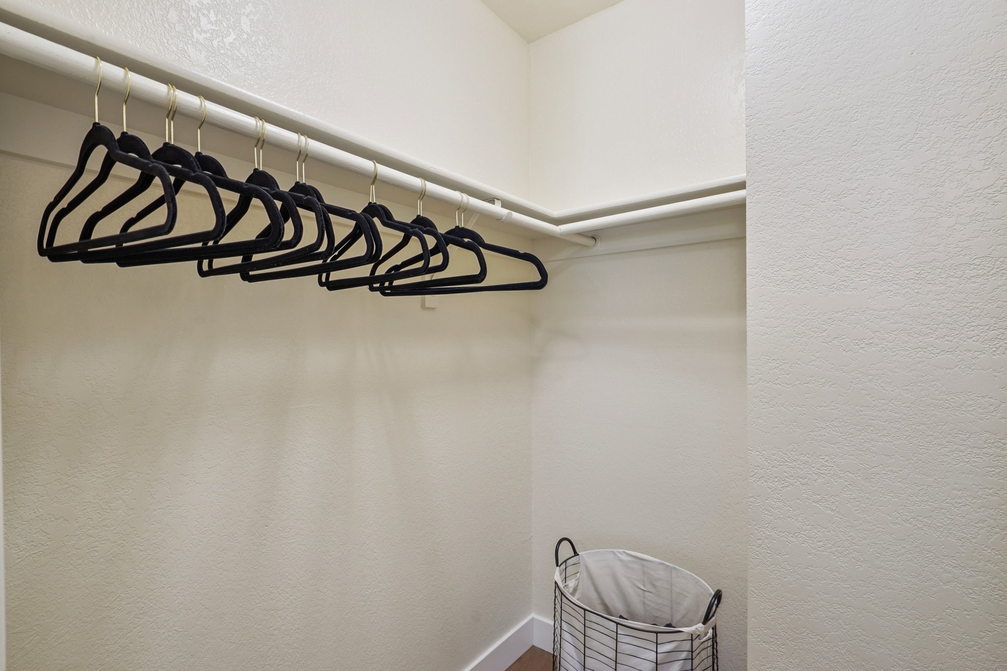 A clean closet with a row of black hangers on a white rod, set against light-colored walls. A wire laundry basket is positioned on the floor, adding to the organized appearance of the space.