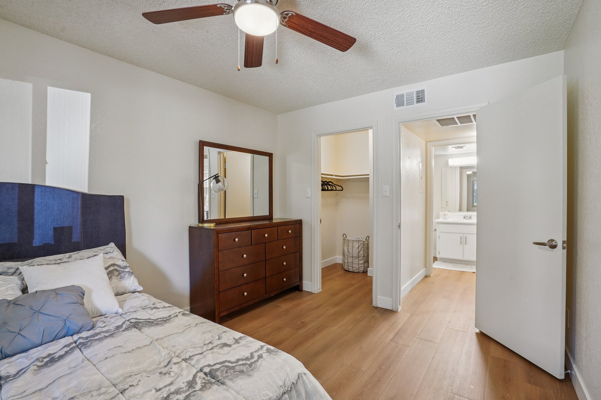 A cozy bedroom featuring a blue upholstered headboard and a wooden dresser with multiple drawers. A ceiling fan is visible above, and a doorway leads to a bathroom area with a mirror and sink. The room has hardwood flooring and neutral-colored walls, creating a warm and inviting atmosphere.