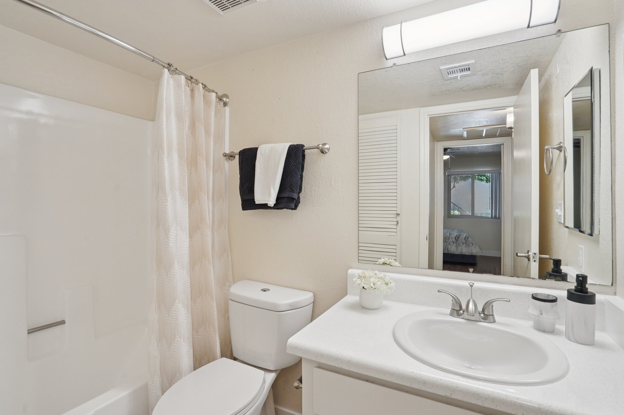 A clean and modern bathroom featuring a white toilet, a sink with a countertop, a mirror, and a shower with a curtain. There are neatly arranged toiletries and a small vase with a flower on the sink. The wall colors are light, contributing to a bright and airy atmosphere. A glimpse of a bedroom is visible through the doorway.