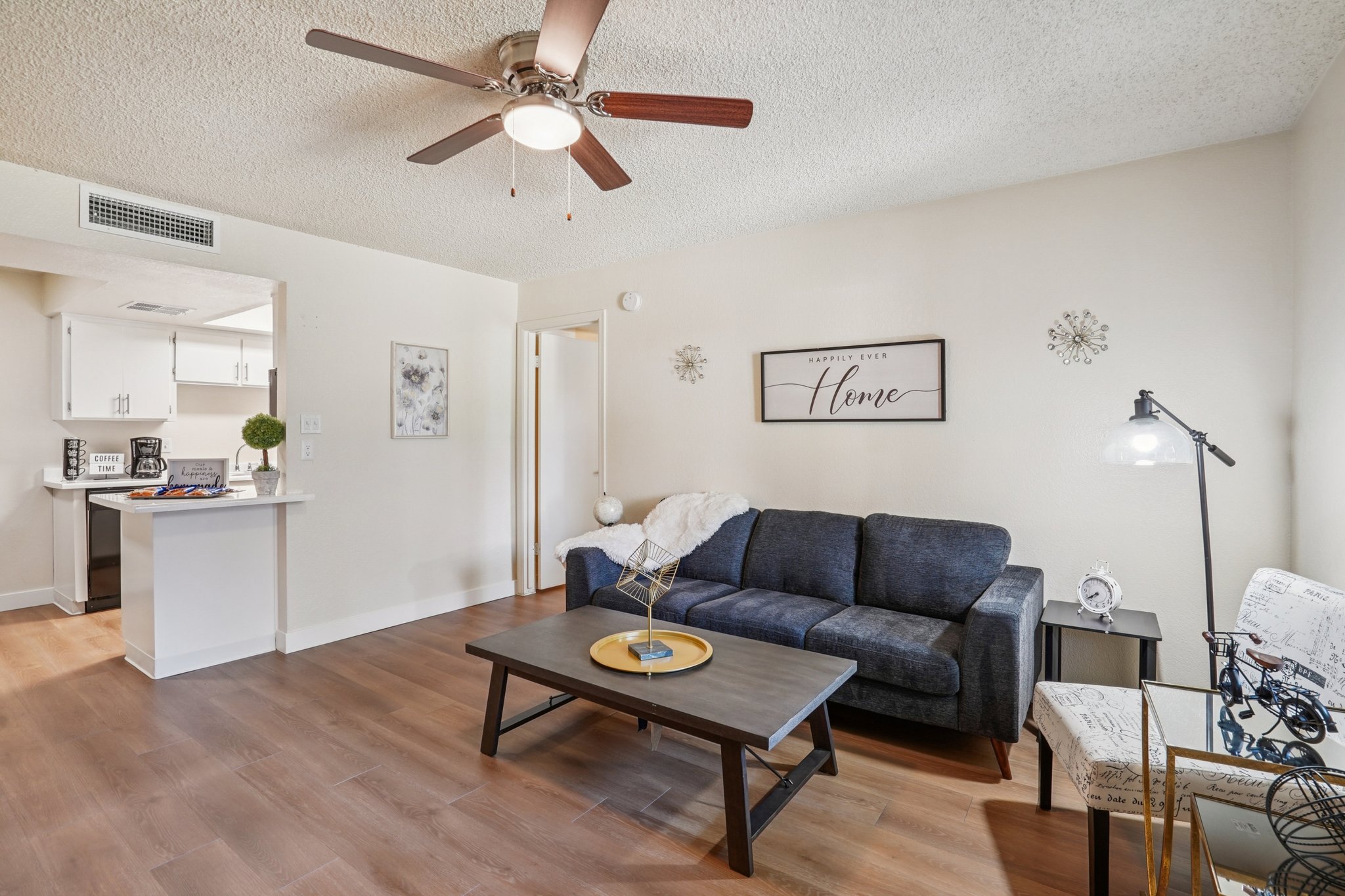 A cozy living room featuring a dark blue sofa, a wooden coffee table, and a decorative lamp. The room has a ceiling fan and light-colored walls, with a framed sign that reads "Home." A small dining area is visible in the background, along with modern kitchen appliances.