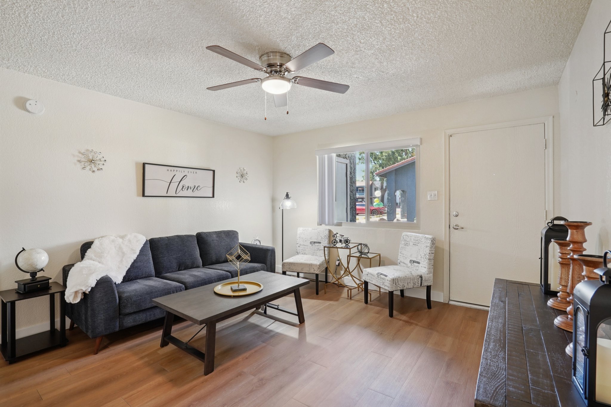 Cozy living room with a light-colored wall and wooden flooring. A dark sofa with a white throw blanket is positioned in front of a wooden coffee table. Two white chairs with decorative patterns are nearby. A fan hangs from the ceiling, and a small decorative globe and a picture frame with "Home" are visible. A door leads outside.