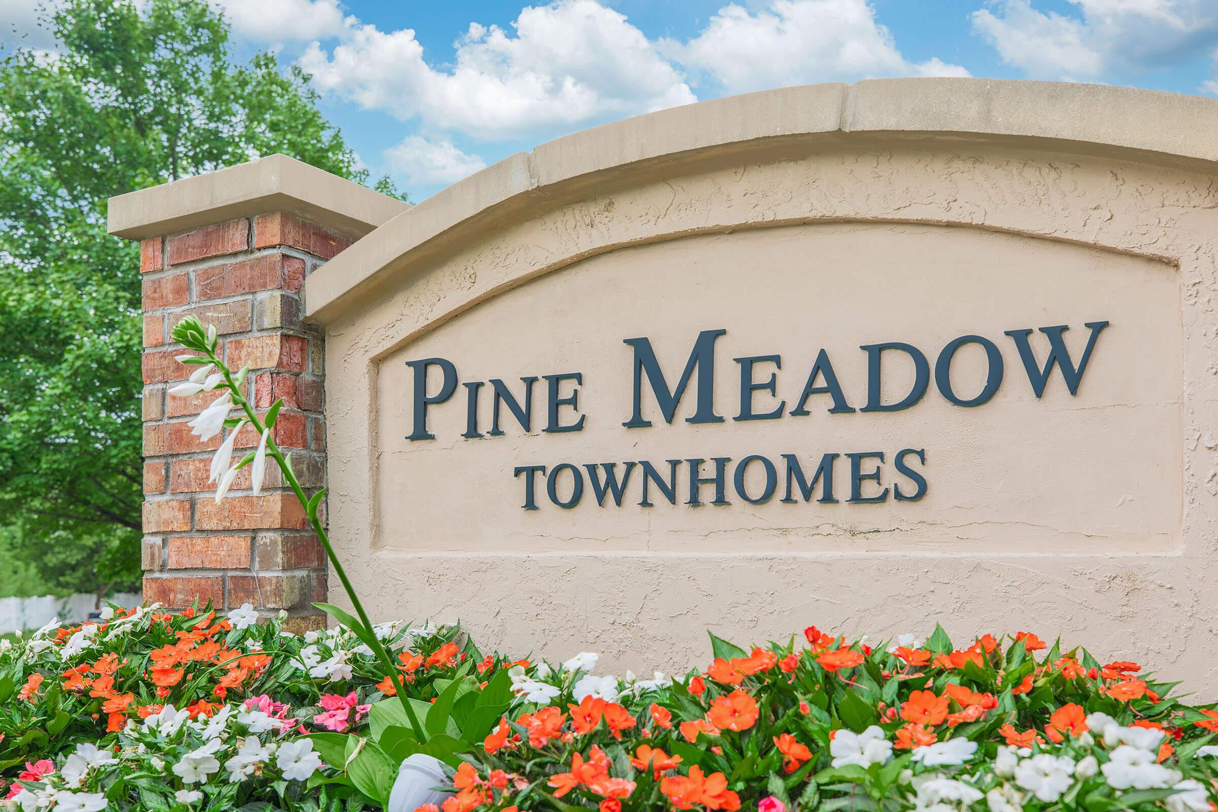 Sign for Pine Meadow Townhomes, featuring the name prominently displayed against a light-colored background. The sign is surrounded by vibrant flowers in shades of orange and white, with greenery in the background and a blue sky with clouds above.