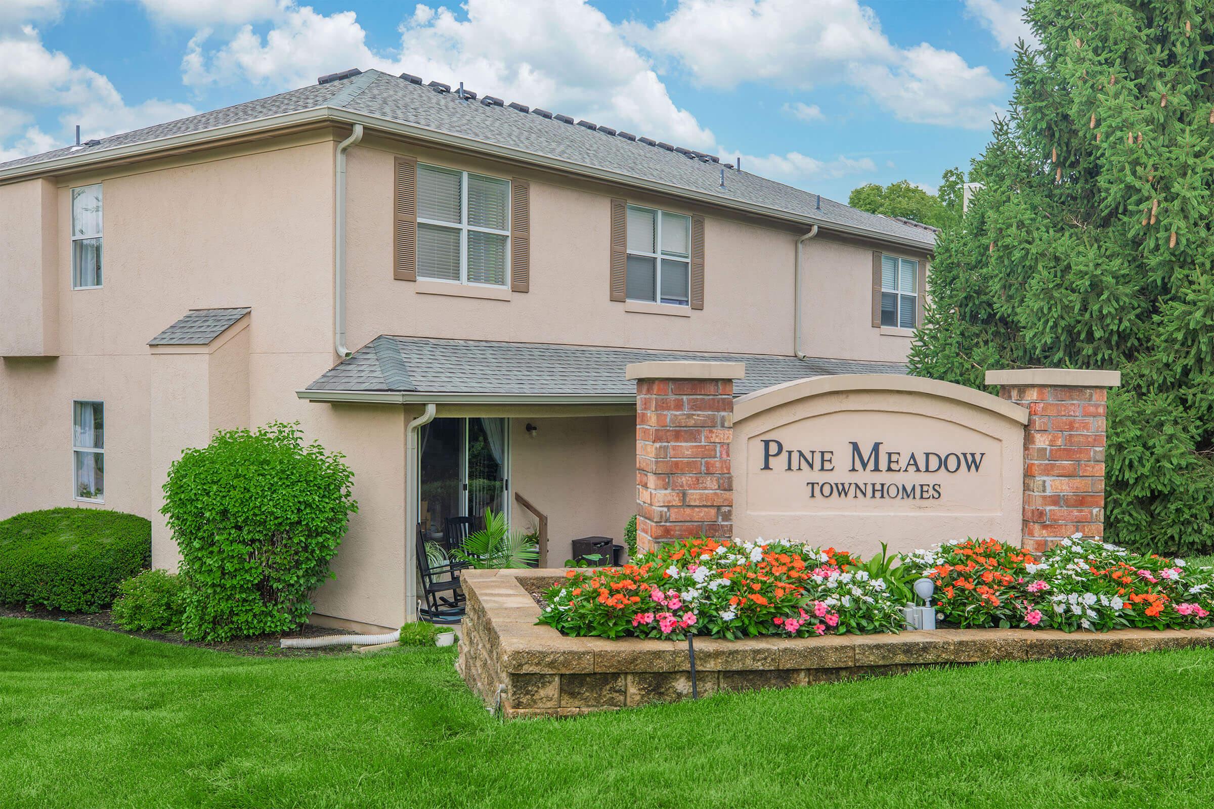 A welcoming sign for Pine Meadow Townhomes, featuring the name prominently displayed on a brick structure. Surrounding the sign are vibrant flower beds and well-maintained greenery, set against a beautiful residential building with multiple windows under a clear blue sky.