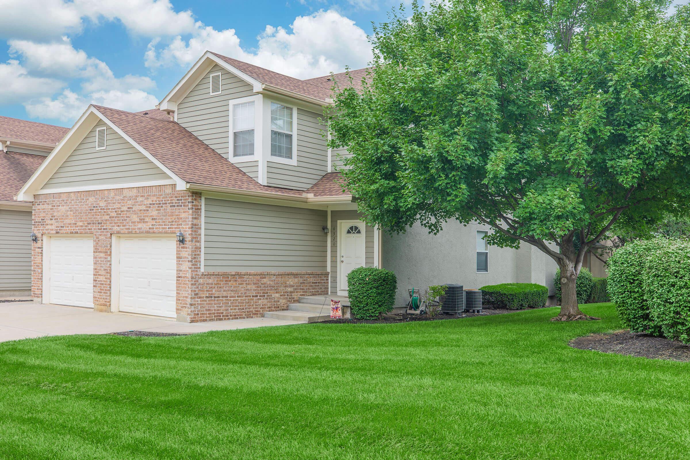 A modern two-story house with a brick facade and grey siding, featuring a well-manicured lawn and a large tree. The driveway leads to a double garage, and the front entryway is visible, adding to the home's curb appeal. The sky is partly cloudy, enhancing the overall bright atmosphere.