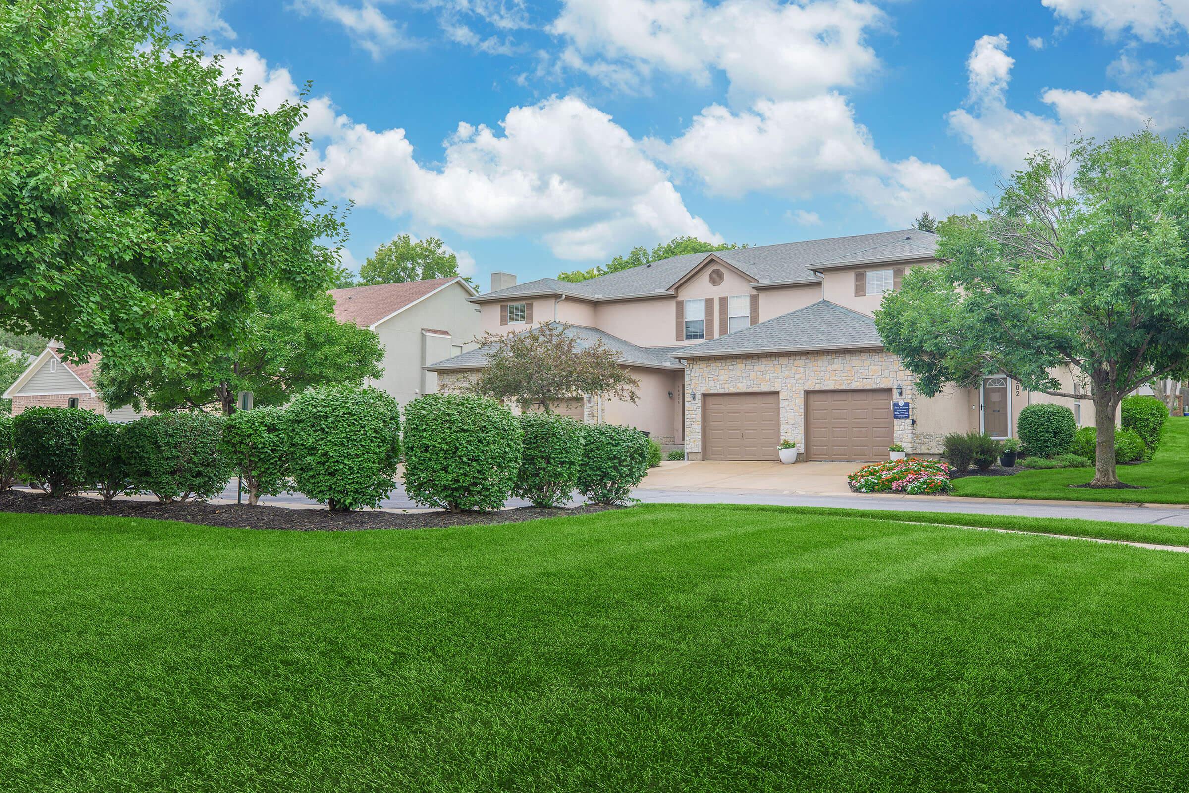 A well-maintained residential property featuring a large lawn, trimmed hedges, and colorful flower beds. The house has a stone and stucco exterior, with a double garage and trees lining the driveway, under a bright blue sky with fluffy white clouds.