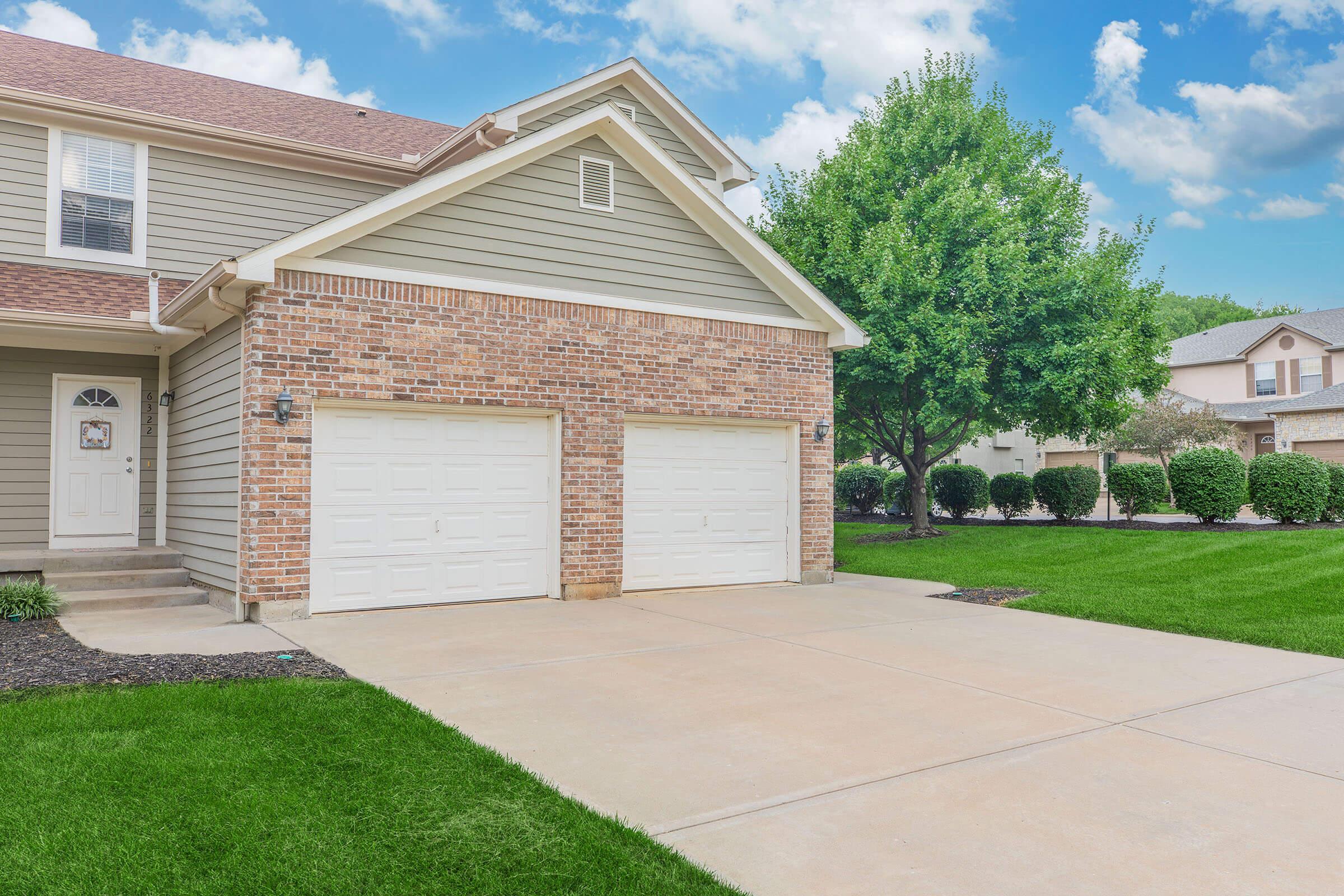 A residential home featuring a two-car garage with white doors, beige siding, and a well-manicured lawn. The driveway is clean and leads to the garage, while green shrubbery and a tree provide a natural backdrop under a partly cloudy sky.