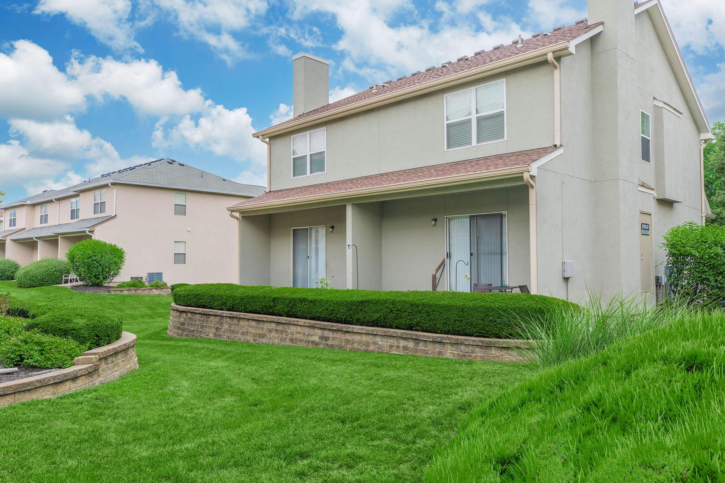 A view of two residential houses in a suburban neighborhood, featuring well-maintained green lawns and hedges. The house in the foreground has a beige exterior, a sloped roof, and sliding glass doors. The sky is partly cloudy, adding a bright backdrop to the scene.