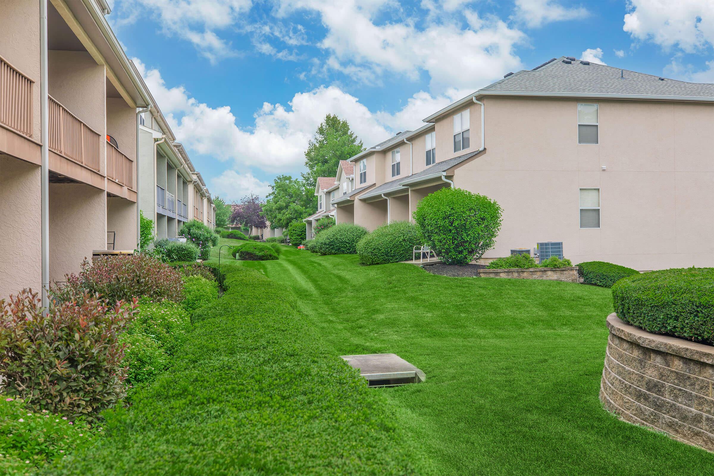A landscaped pathway lined with neatly trimmed shrubs and grass, flanked by residential buildings on either side. The sky above is bright and partly cloudy, creating a serene and inviting atmosphere. The well-maintained green space invites leisurely walks or relaxation.