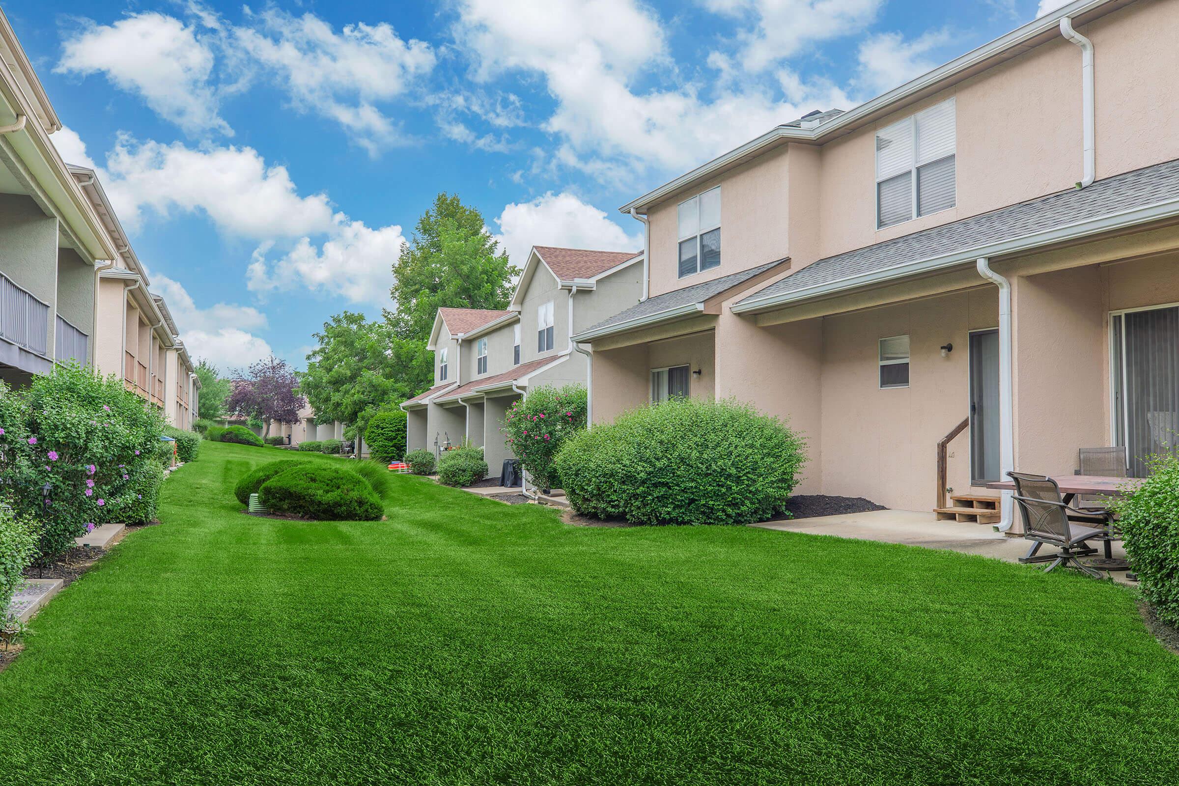 A view of a well-maintained residential area featuring two-story homes with light-colored exteriors, surrounded by lush green lawns and shrubs. A pathway runs through the manicured grass, leading to a small seating area on a patio. The sky is bright and partly cloudy, adding to the serene atmosphere.