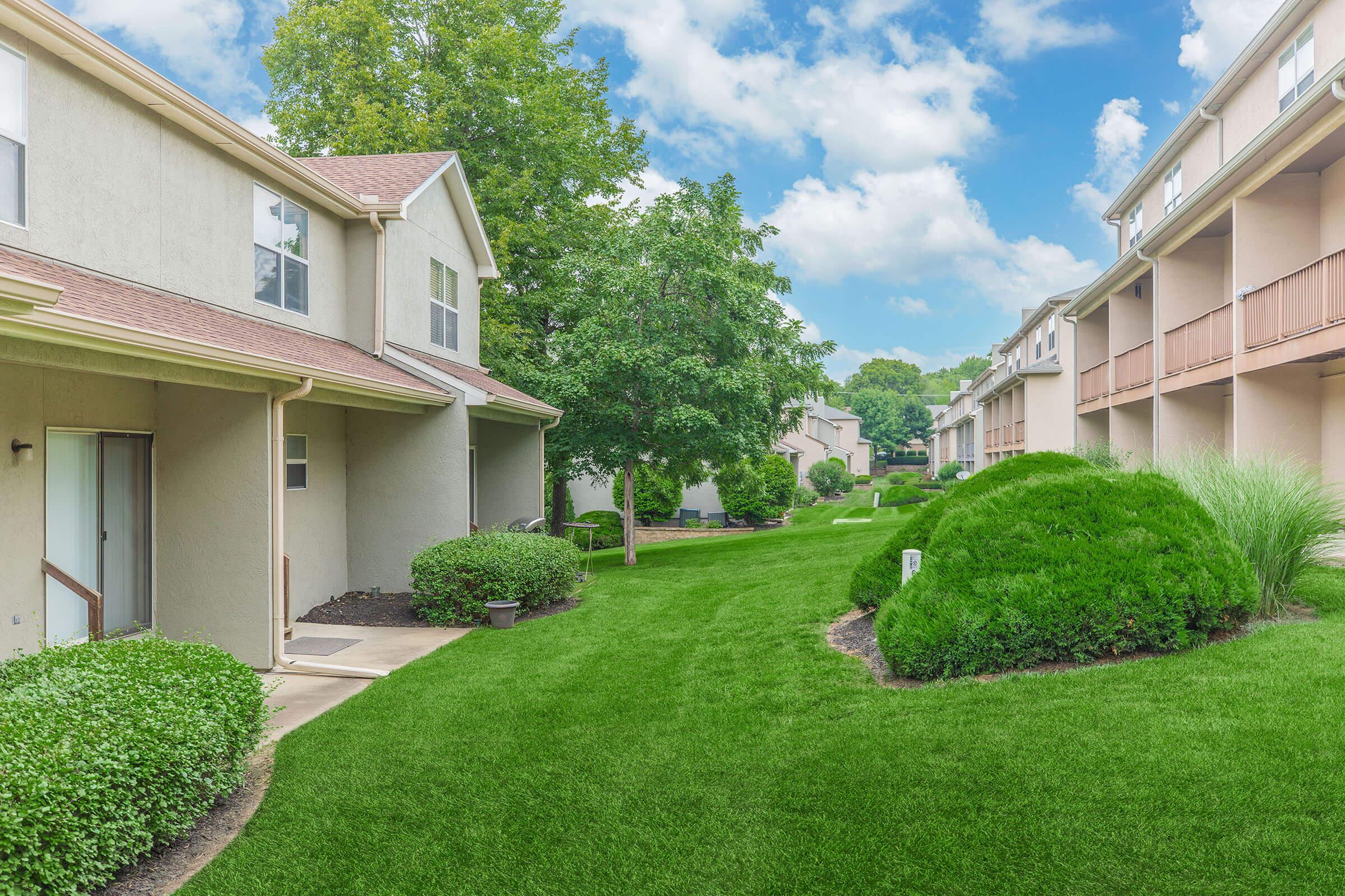 A well-maintained residential area featuring two-story buildings with green lawns, shrubs, and small trees. The scene is sunny with a bright blue sky and fluffy white clouds, creating a peaceful, inviting atmosphere. The walkways are lined with grass, enhancing the tranquil environment.