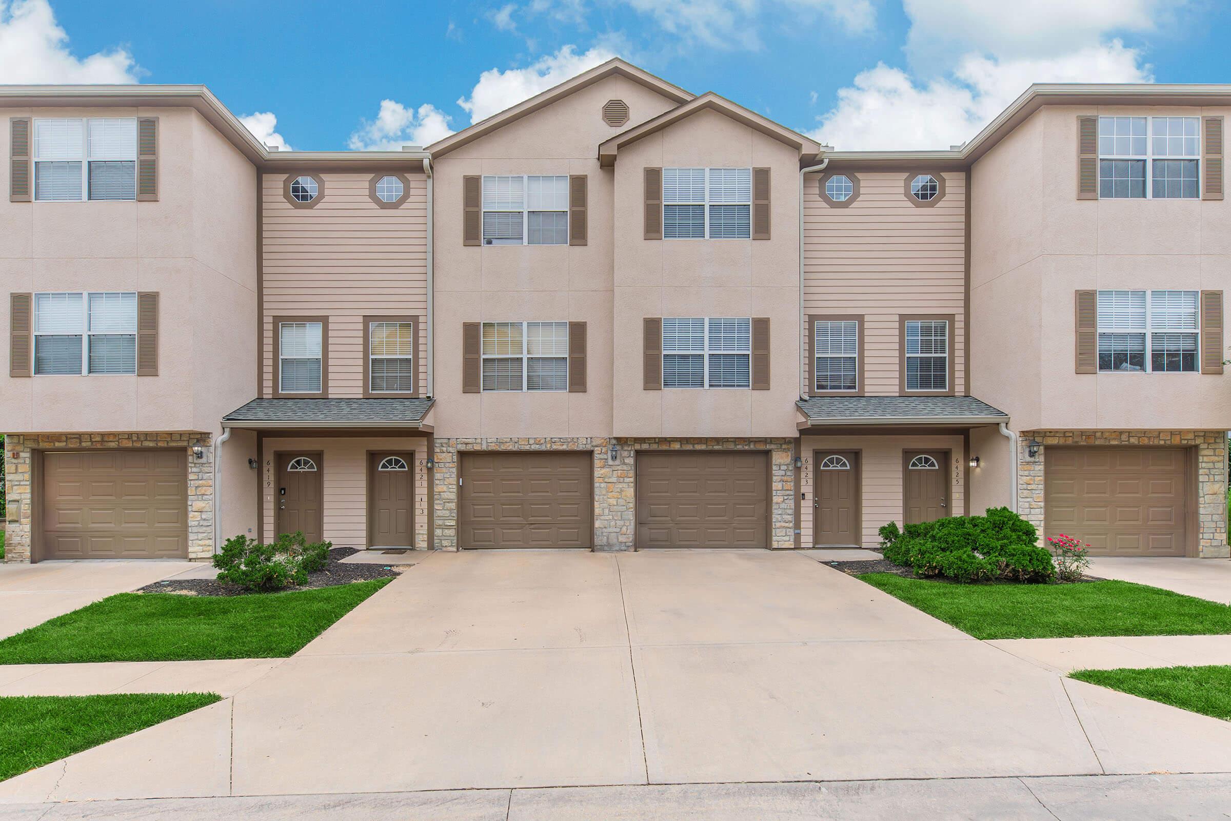 A row of residential townhomes with tan siding and stone accents. Each unit features a garage door and windows with white frames. The pathway in front is paved, with green grass and small bushes on either side, set against a blue sky with a few clouds.