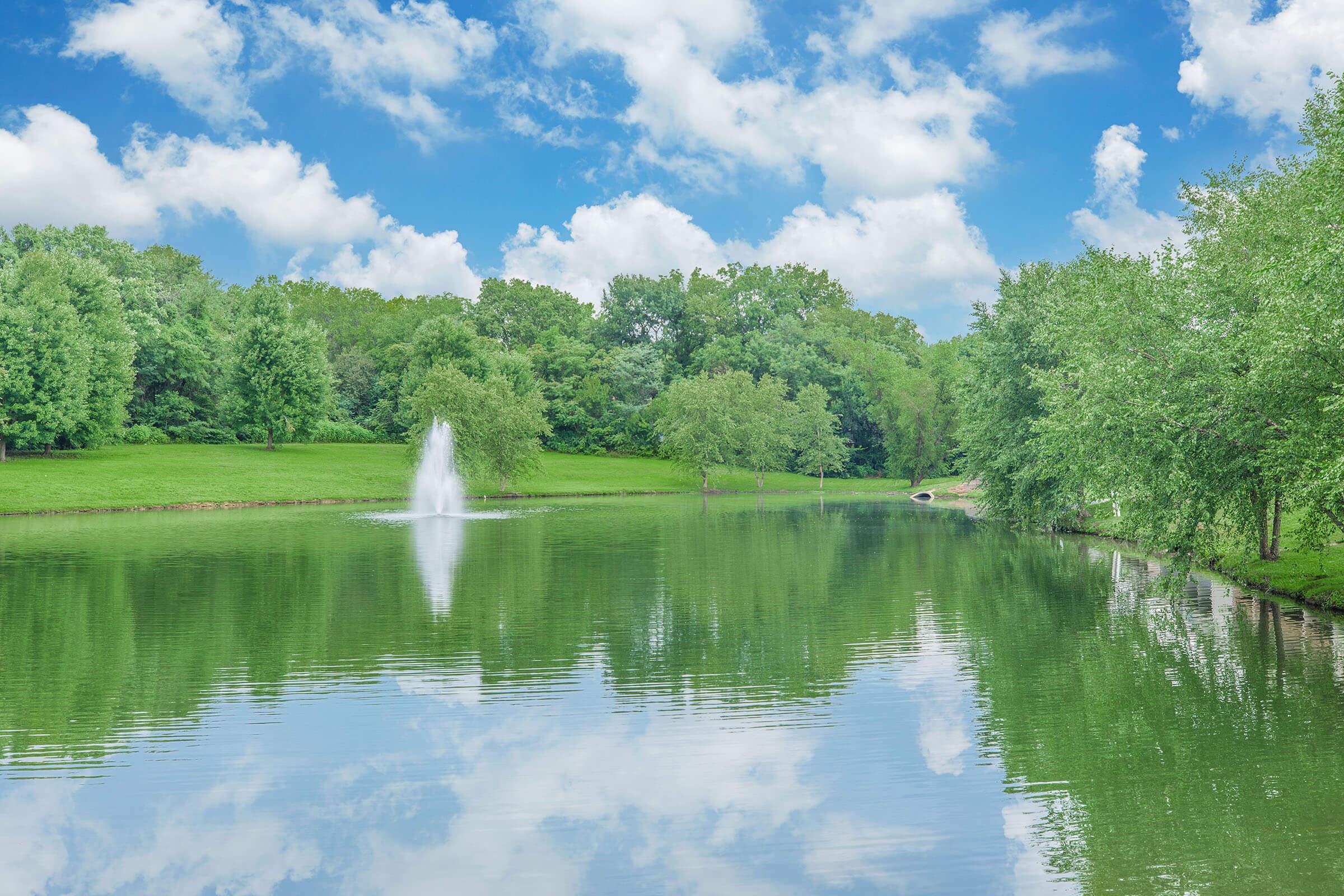 A serene lake surrounded by lush green trees under a partly cloudy sky. A fountain sprays water in the middle of the lake, creating ripples on the reflective surface. The tranquil scene conveys a sense of peace and natural beauty.