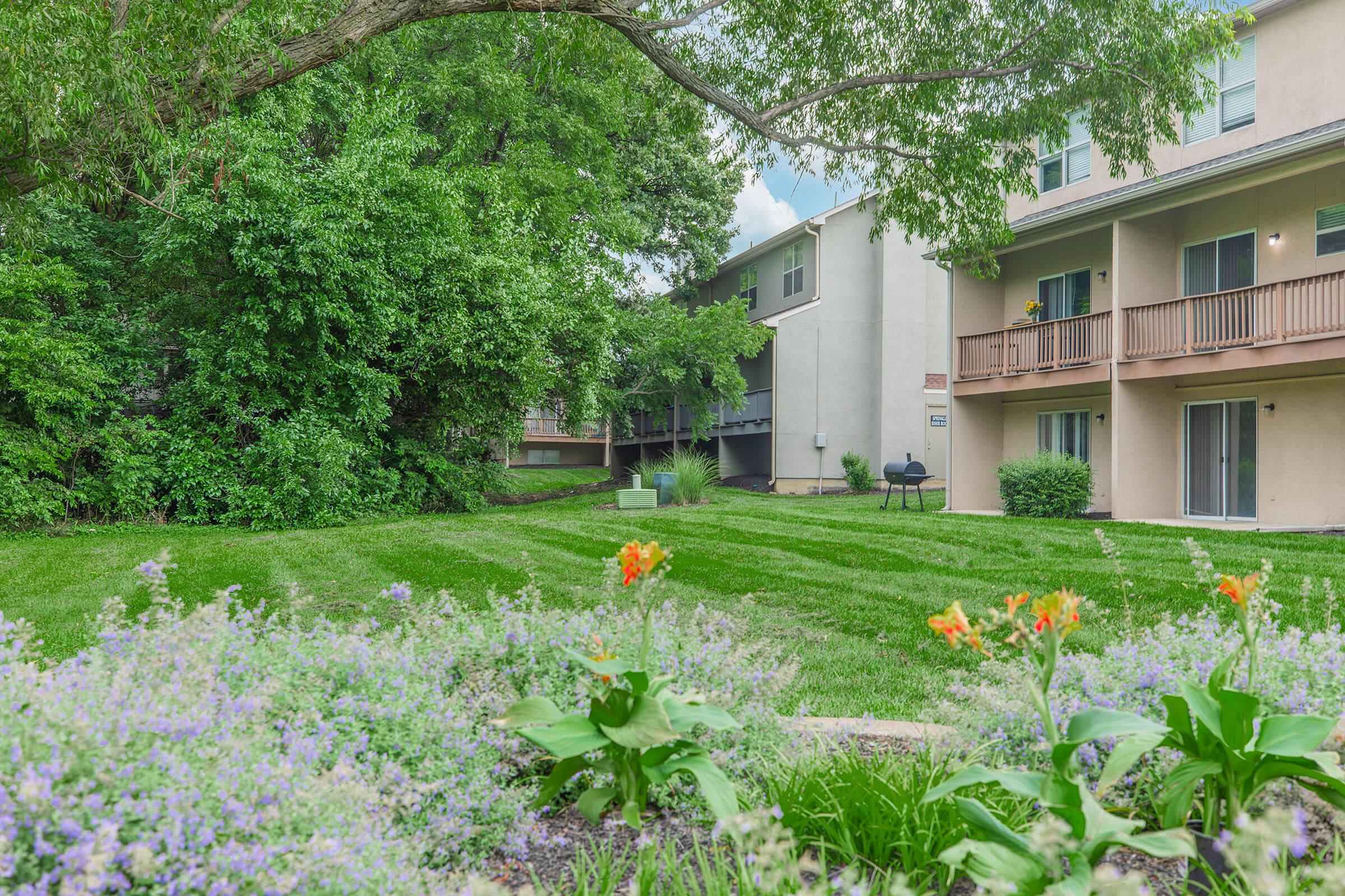 A landscaped area featuring well-maintained green grass and flower beds with orange flowers. In the background, two residential buildings with balconies are visible, surrounded by lush trees and shrubs. A small grill is placed on the patio of one building. The sky is partly cloudy, creating a bright atmosphere.