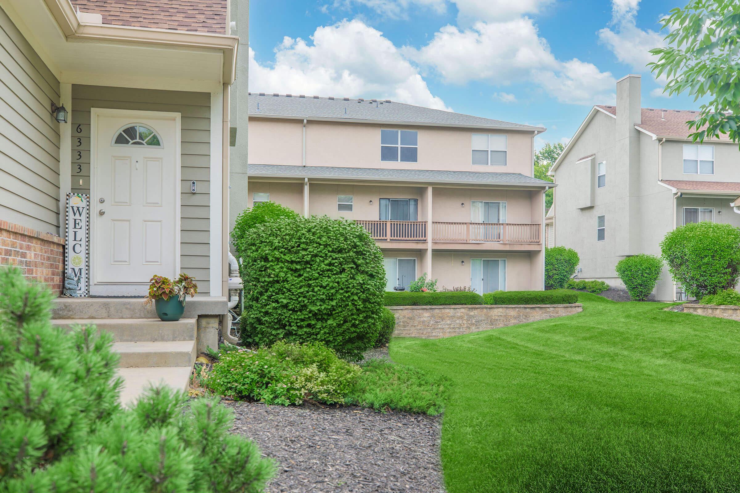 A well-maintained residential area featuring two apartment buildings. The foreground includes a pathway leading to a front door adorned with a welcome sign, surrounded by lush greenery and manicured lawns. The sky is bright with fluffy clouds, enhancing the inviting atmosphere of the neighborhood.