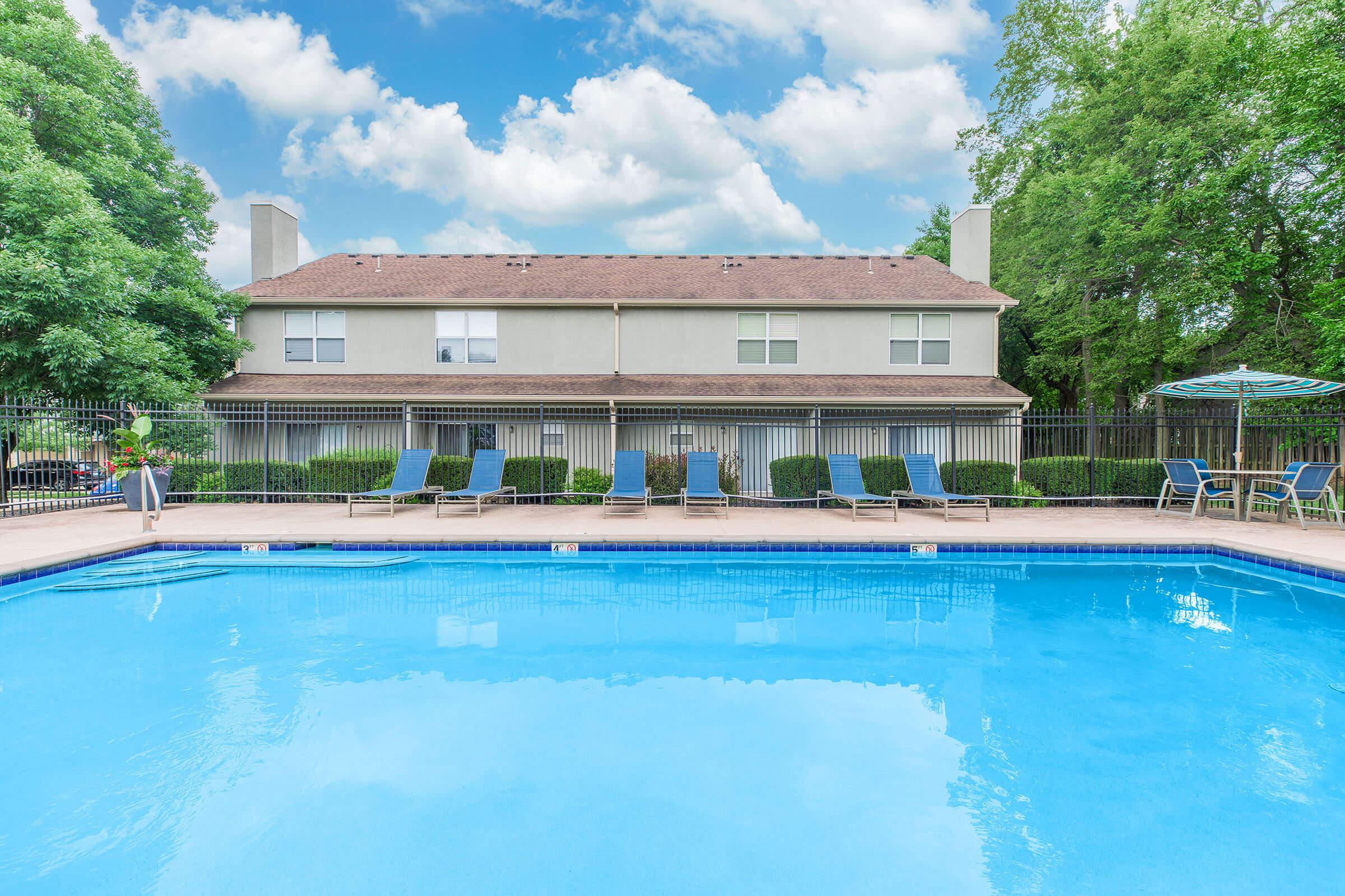 A clear blue swimming pool in the foreground, surrounded by lounge chairs and a shaded area with an umbrella. In the background, a modern residential building with large windows and green trees lining the property under a bright blue sky with fluffy white clouds.