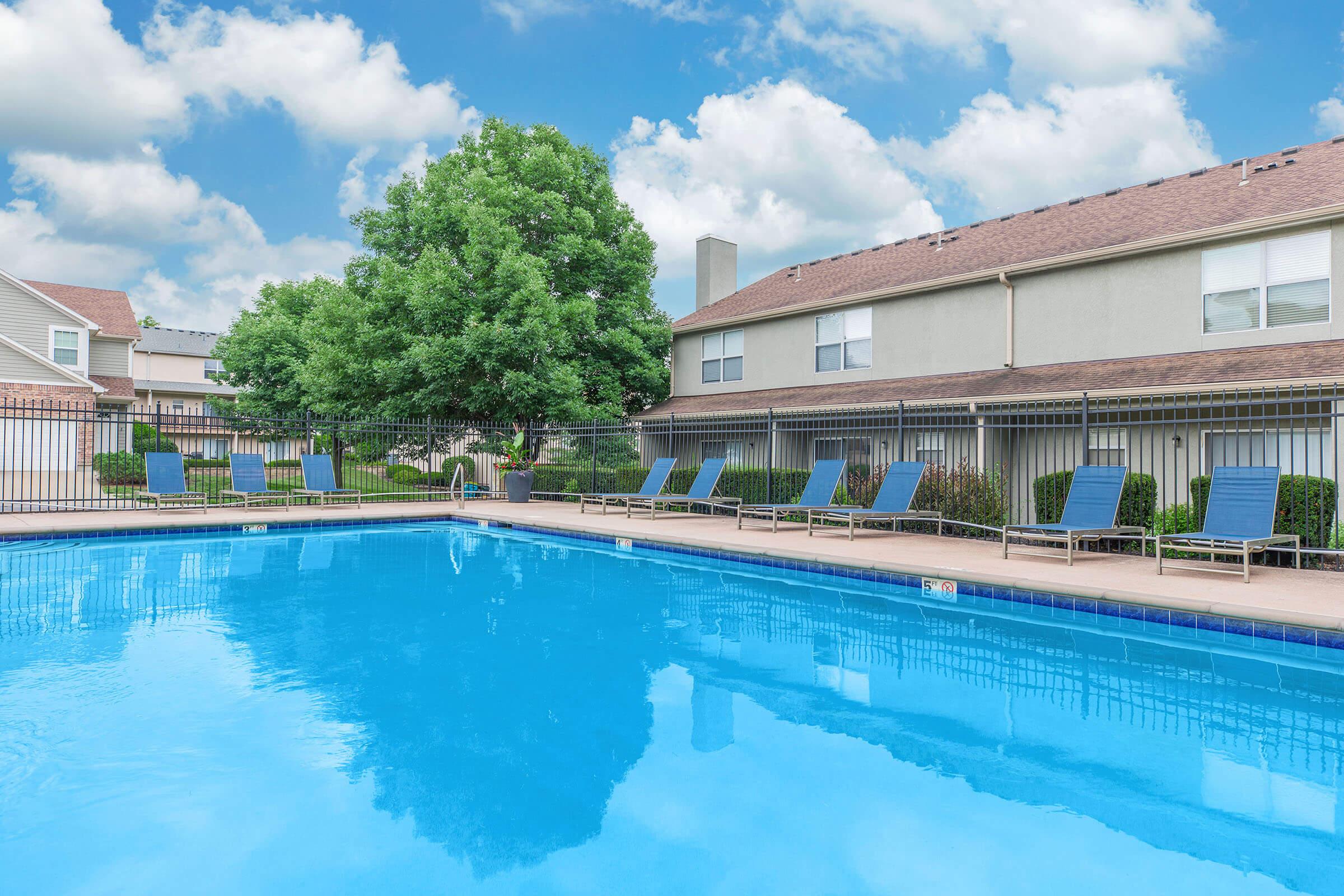 A clear blue swimming pool surrounded by lounge chairs sits in a well-maintained outdoor area. Green trees and manicured bushes line the poolside, with two-story buildings visible in the background under a partly cloudy sky.