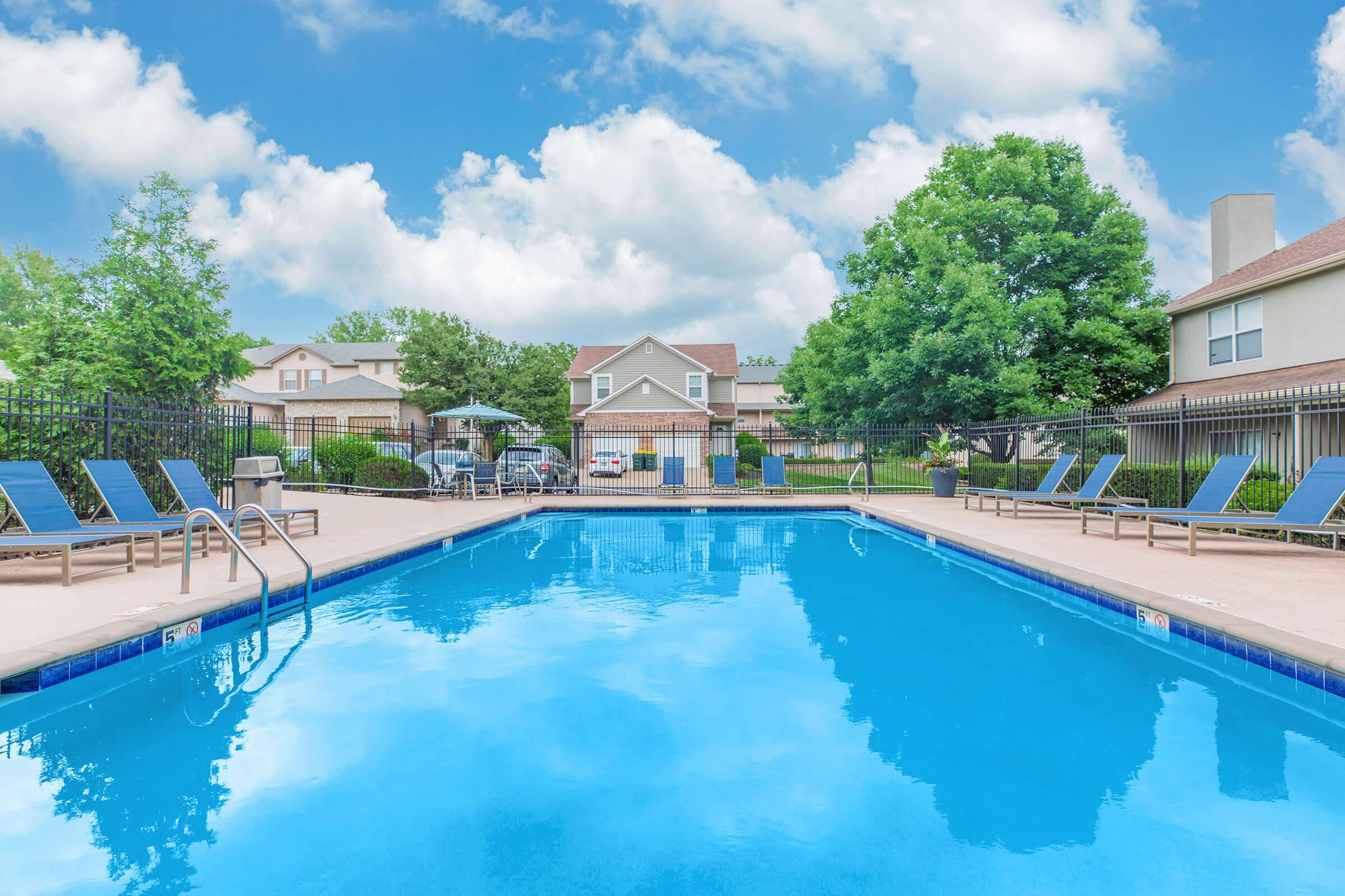 A clear blue swimming pool surrounded by lounge chairs, with green trees and a few residential buildings in the background under a partly cloudy sky. The area is well maintained, creating a relaxing outdoor space.