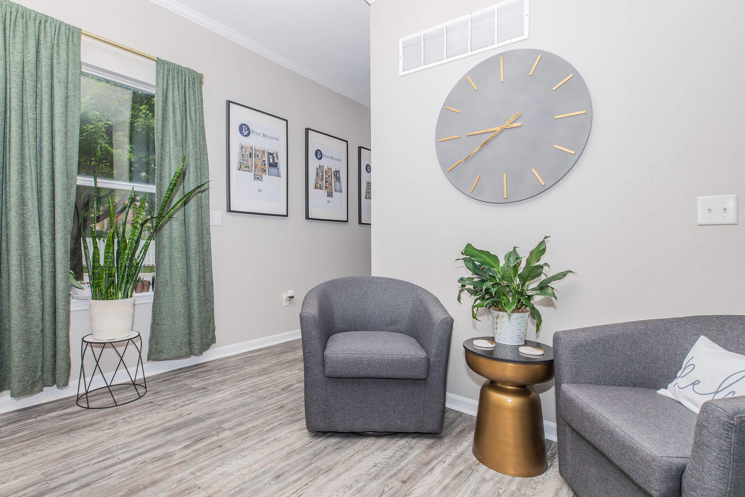 A cozy living space featuring a gray armchair and a round gold table. There’s a tall green plant in a white pot and curtains on the window. Two framed pictures are on the wall, and a large wall clock with a gray face and silver hands is above the table, complementing the modern decor.