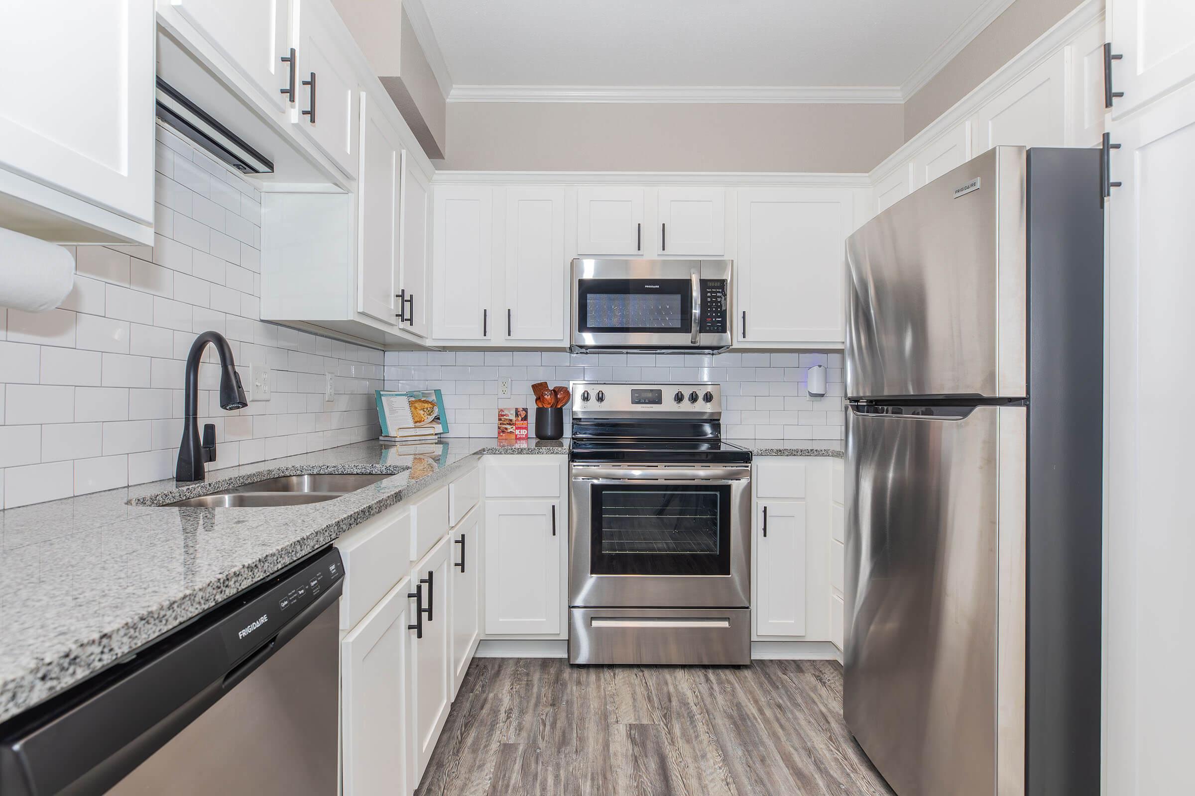 Modern kitchen featuring white cabinetry, stainless steel appliances including a microwave, stove, and refrigerator, granite countertops, and a stylish backsplash. The floor is adorned with laminate planks, and the space is well-lit with neutral wall colors, creating a clean and inviting atmosphere.
