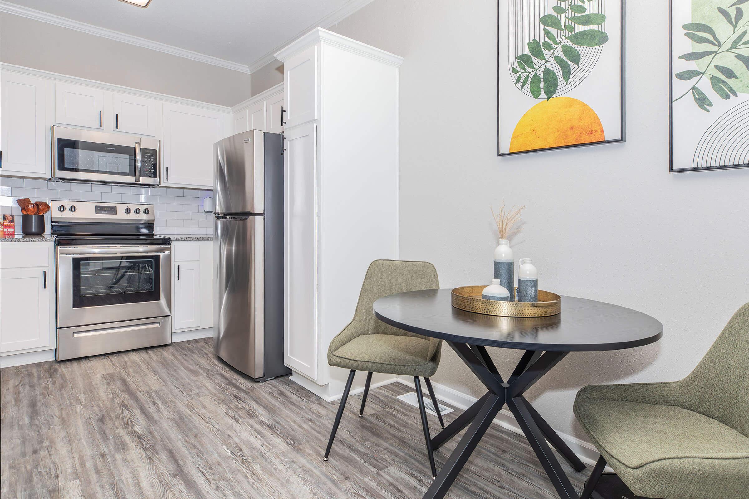 A modern kitchen featuring white cabinets, stainless steel appliances, and a small round black dining table with two green chairs. The walls are decorated with two leaf-patterned prints, and the flooring is a light wood finish. A vase with decorative elements is on the table.