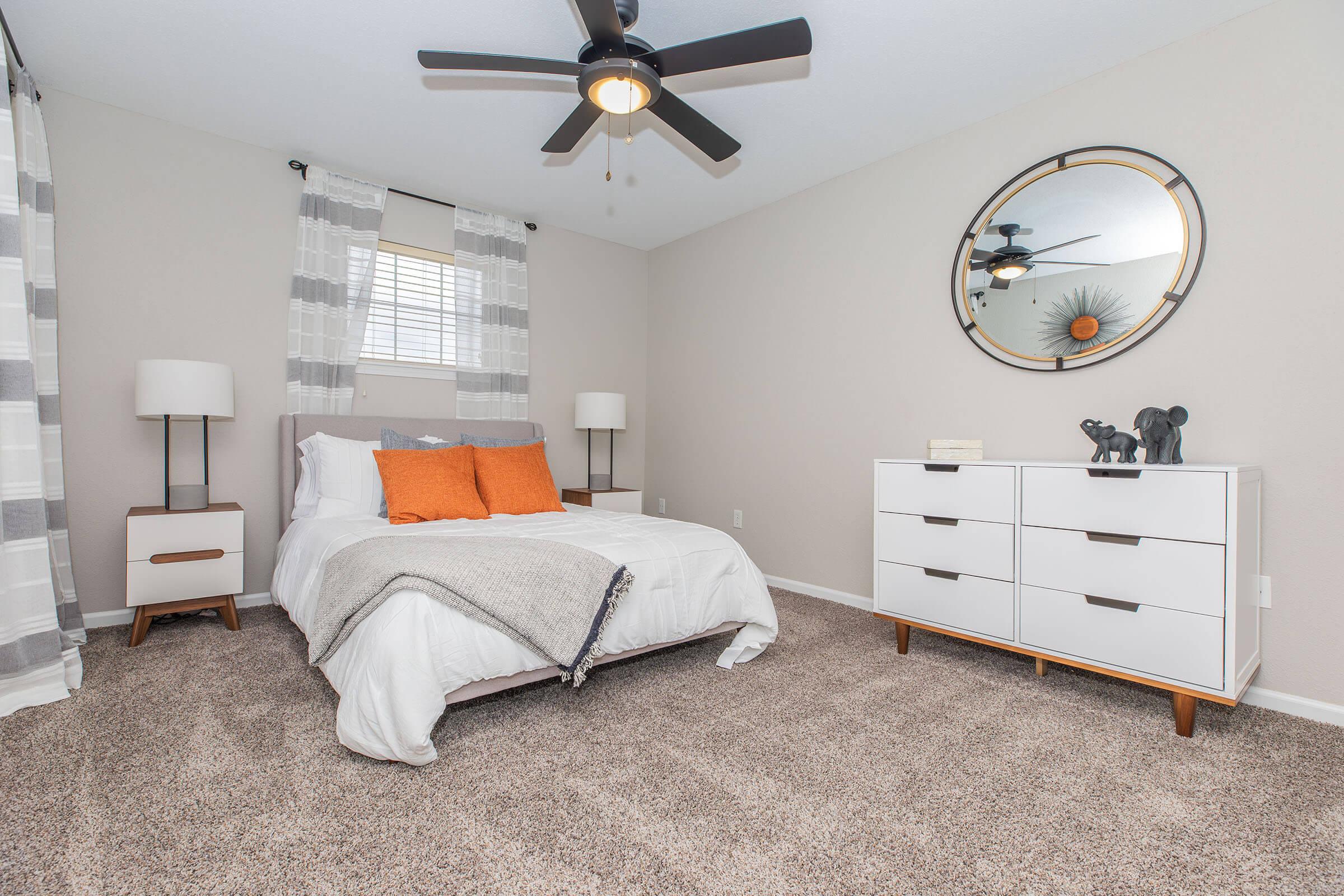 A cozy bedroom featuring a neatly made bed with white linens and orange accent pillows, a stylish dresser, a circular mirror on the wall, and two modern bedside lamps. The room has light gray walls, soft carpeting, and patterned curtains, creating a warm and inviting atmosphere.
