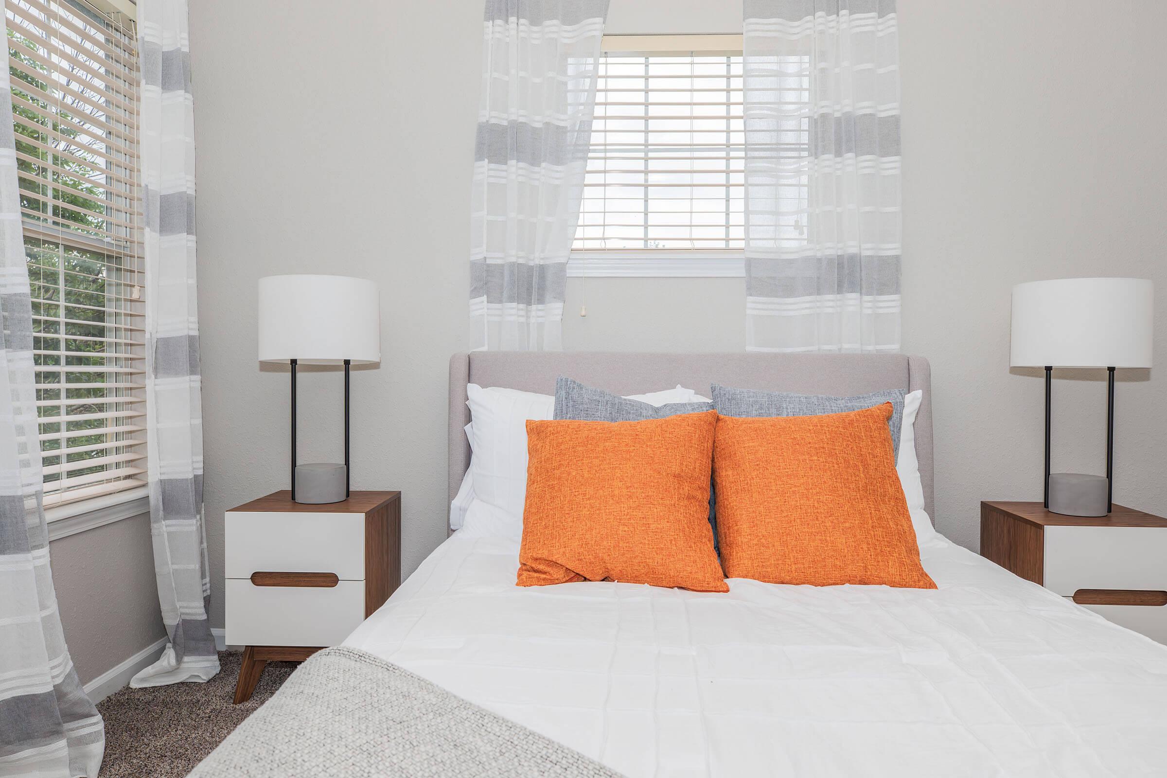 A cozy bedroom featuring a light gray bed with decorative pillows, flanked by bedside tables with modern lamps. Natural light streams in through a window adorned with gray and white curtains. The warm orange pillows add a pop of color to the serene aesthetic.