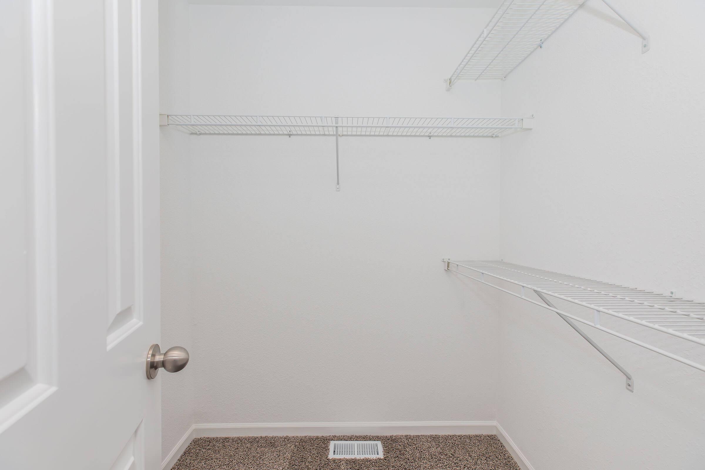 A clean, empty closet with white walls and shelves. The closet features wire shelving along the sides and back, with a carpeted floor. A door with a polished knob is partially visible on the left side.