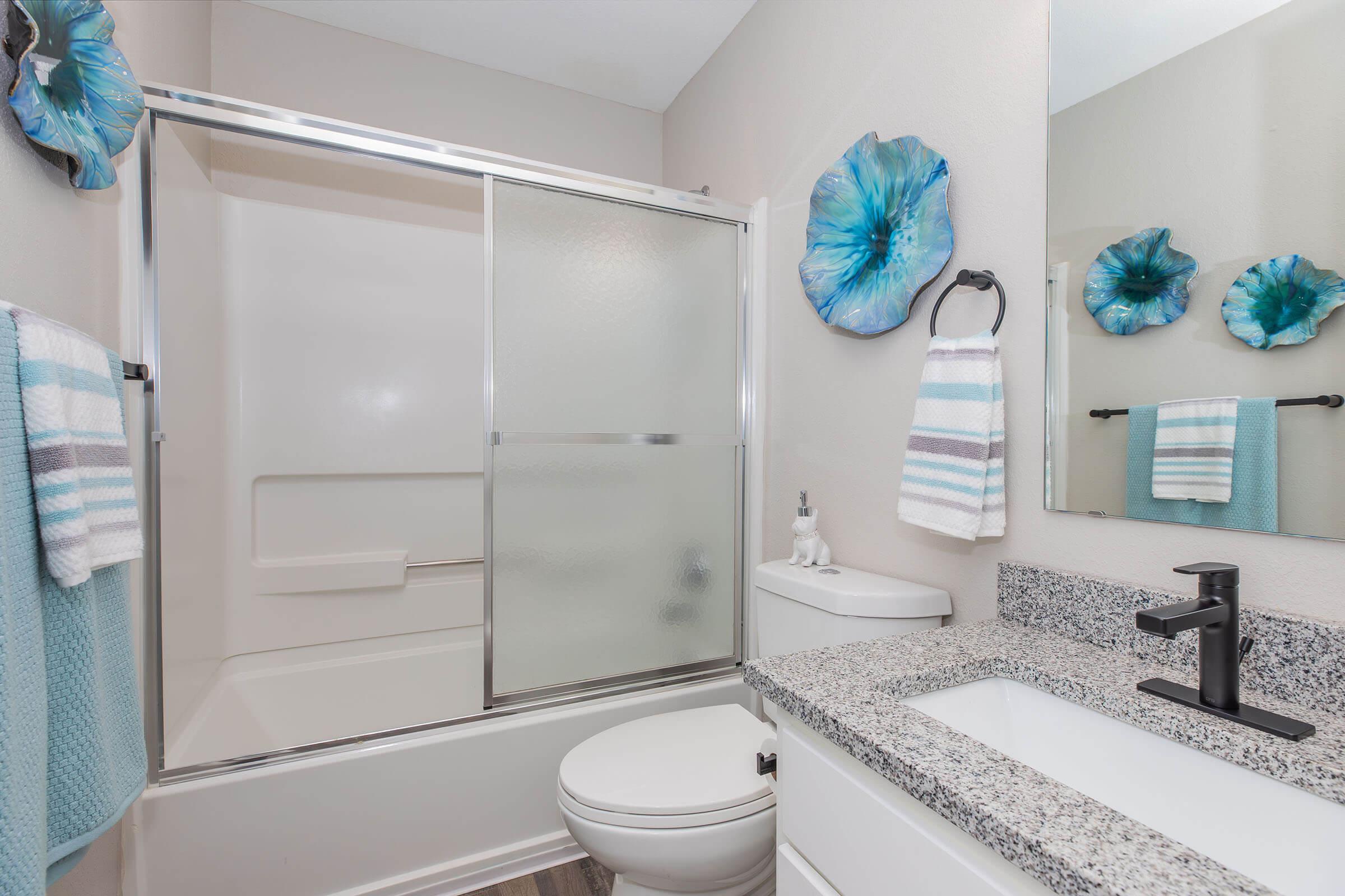 A modern bathroom featuring a glass shower enclosure, a white toilet, and a sleek sink with a granite countertop. The walls are decorated with blue floral wall art and there are light blue and white towels hanging. The space is bright and well-organized, showcasing a clean and contemporary design.