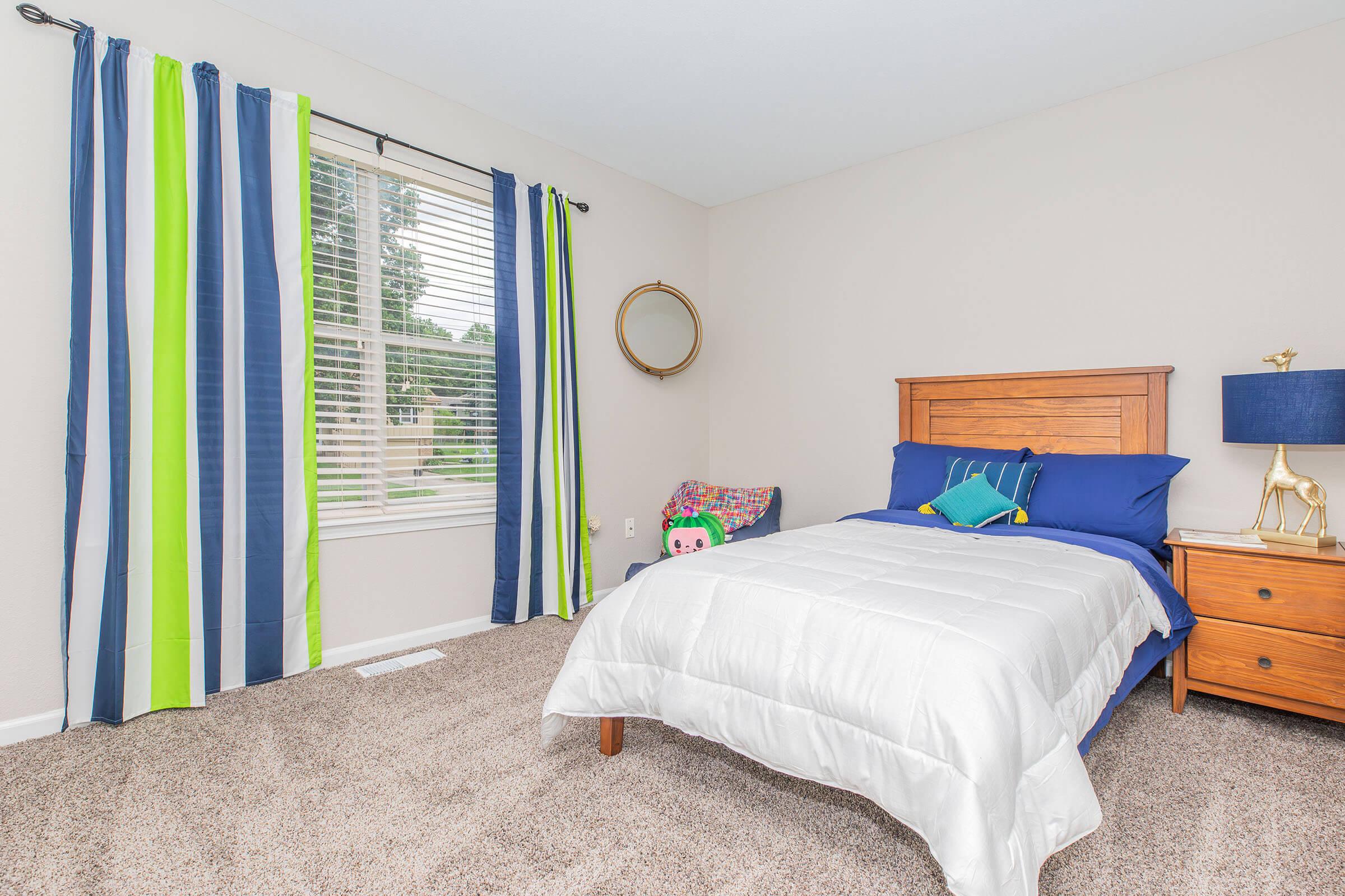 A cozy bedroom featuring a wooden bed with a white comforter, blue and green accent pillows. A nightstand with a blue lamp and a decorative item sits beside the bed. Large windows dressed with striped curtains allow natural light in, illuminating the soft carpeted floor.