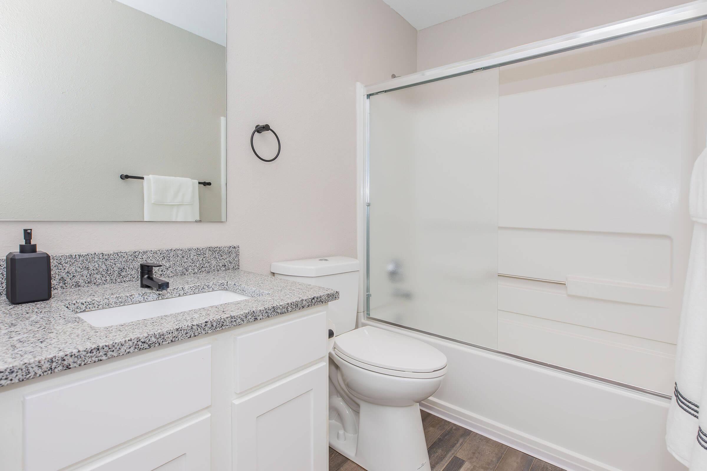 A clean and modern bathroom featuring a white toilet, a glass shower enclosure, a granite countertop sink with a black faucet, and a wall-mounted mirror. There is a towel hanging on a rack and a black soap dispenser on the countertop, with light-colored walls and vinyl flooring.