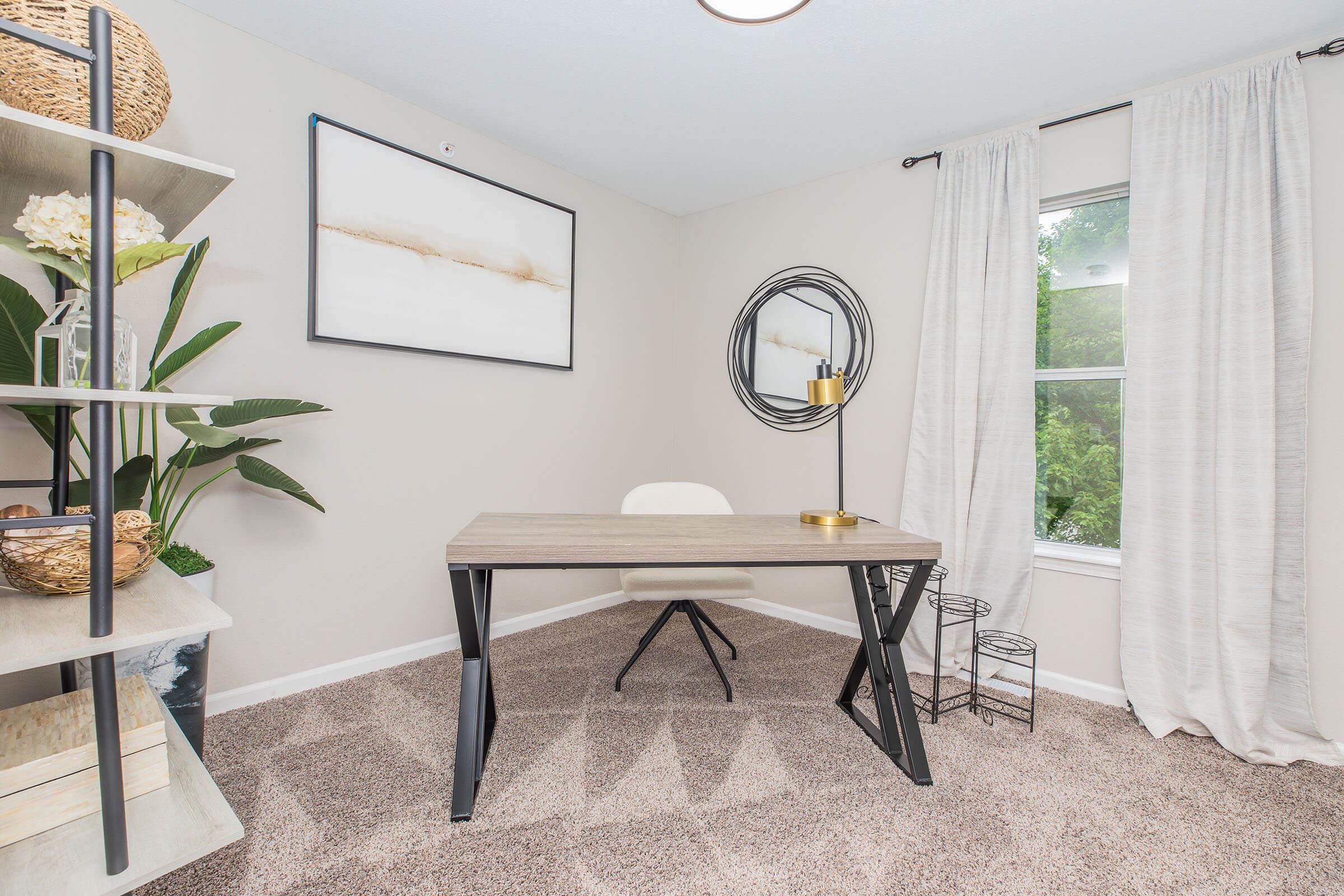 A modern home office featuring a minimalist desk with a white chair, a round mirror on the wall, and a large artwork piece. A shelf displays decorative plants and items. The room is well-lit with natural light coming through a window, complemented by light-colored curtains and a cozy carpeted floor.