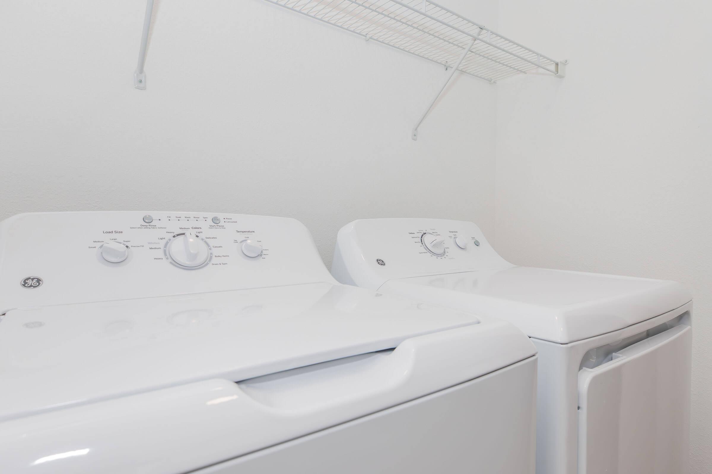 Two white laundry appliances, a washing machine on the left and a dryer on the right, positioned side by side in a clean, bright indoor laundry space. Above them is a metallic wire shelf attached to the wall. The room has a neutral-colored wall and ample lighting.