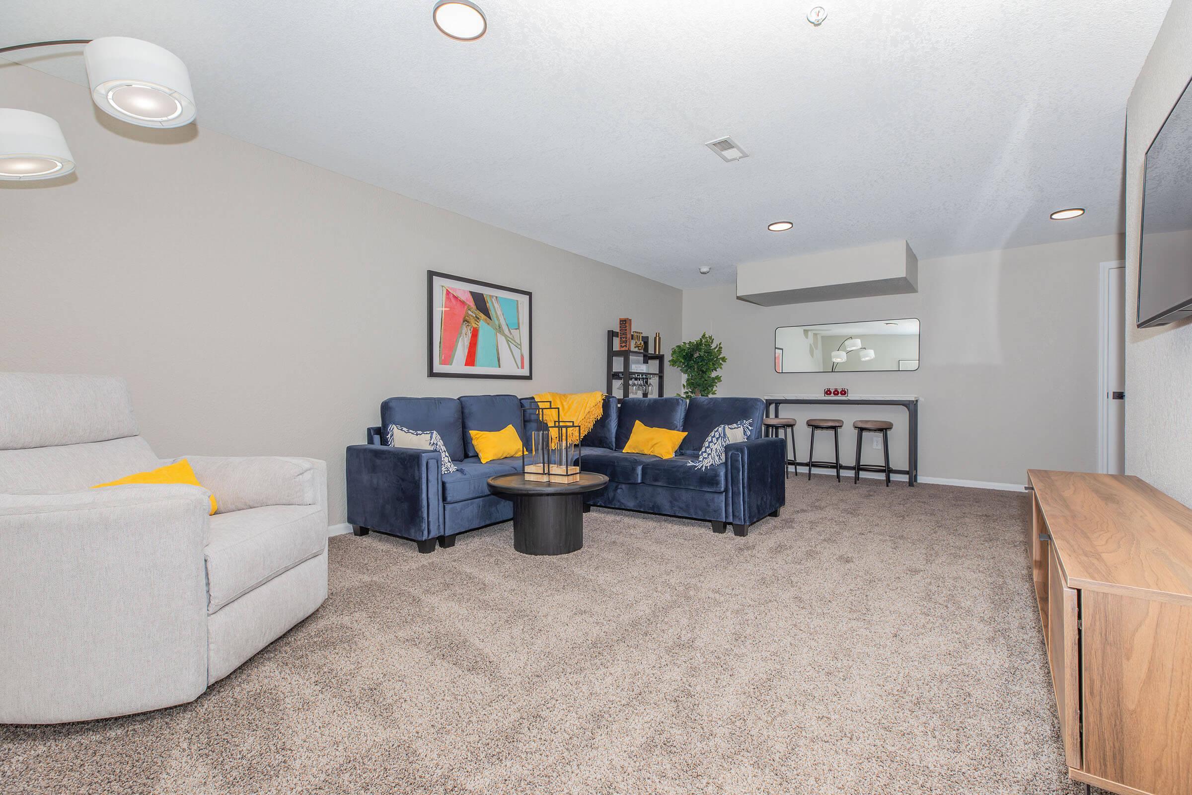 A cozy living room featuring two blue sofas with yellow pillows, a round coffee table, and a modern light fixture. There is a beige carpet, a small bar area with stools in the background, and a colorful artwork on the wall. A plant adds a touch of greenery to the space.