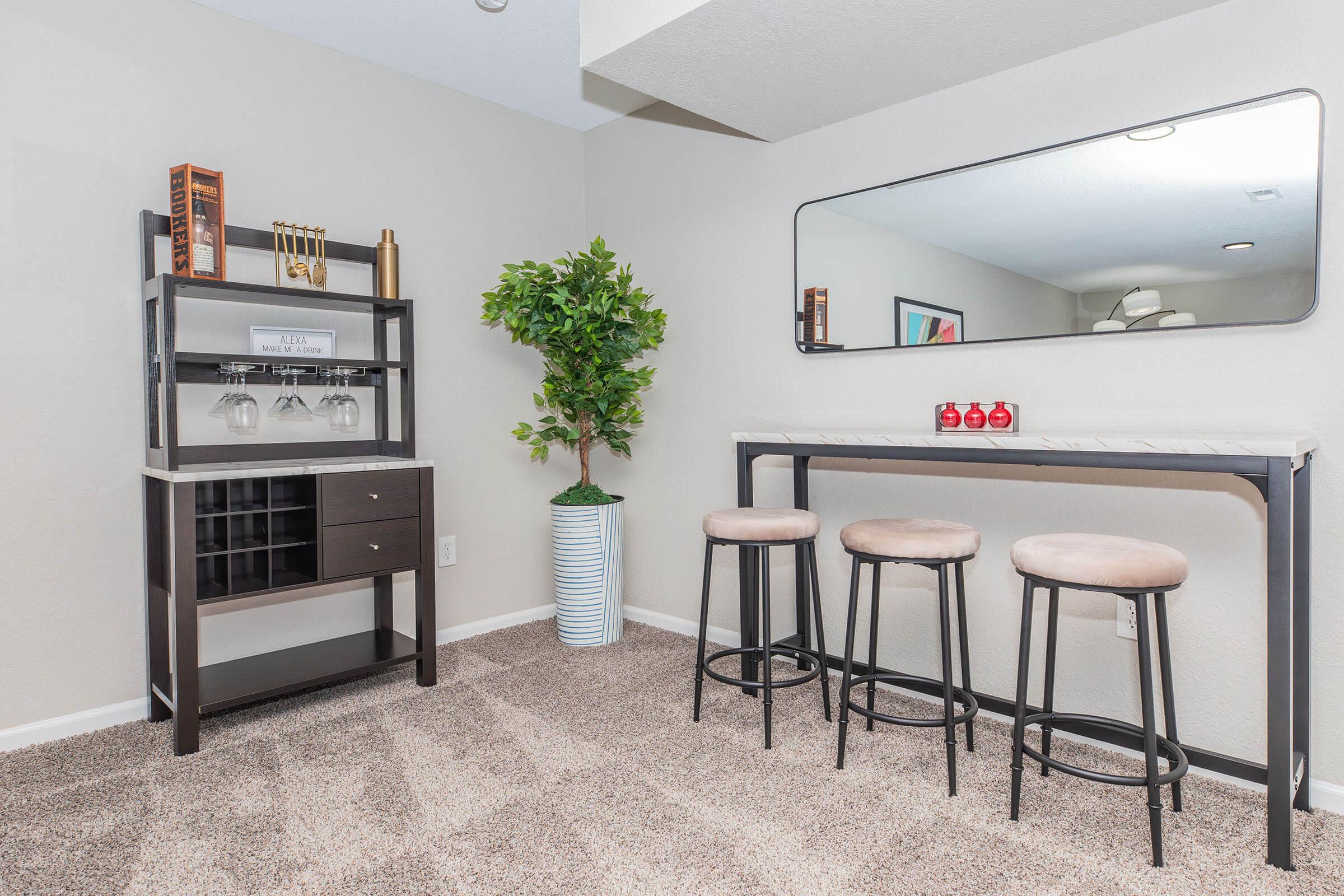 A cozy interior space featuring a modern bar area with a black shelving unit holding glasses, a decorative plant in a blue striped pot, and a long table with three stylish stools. A large mirror hangs above the table, and soft carpeting covers the floor, creating an inviting atmosphere.