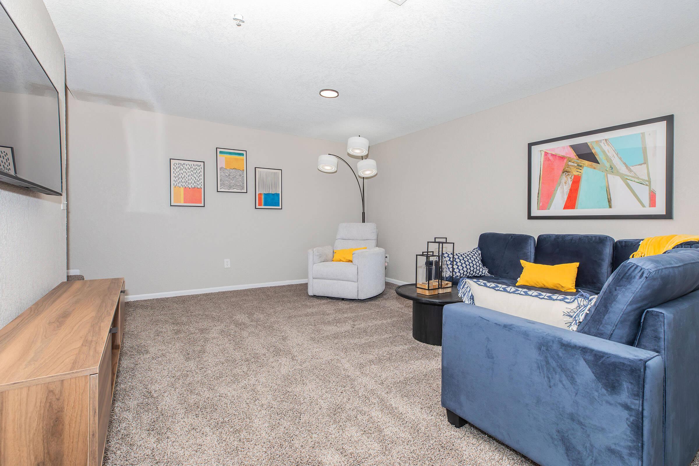 A cozy living room featuring a blue sofa with yellow pillows, a light gray armchair, a round coffee table, and a wooden TV stand. The walls are adorned with colorful framed artwork. Bright, modern lighting enhances the relaxed atmosphere, complemented by soft, beige carpet.