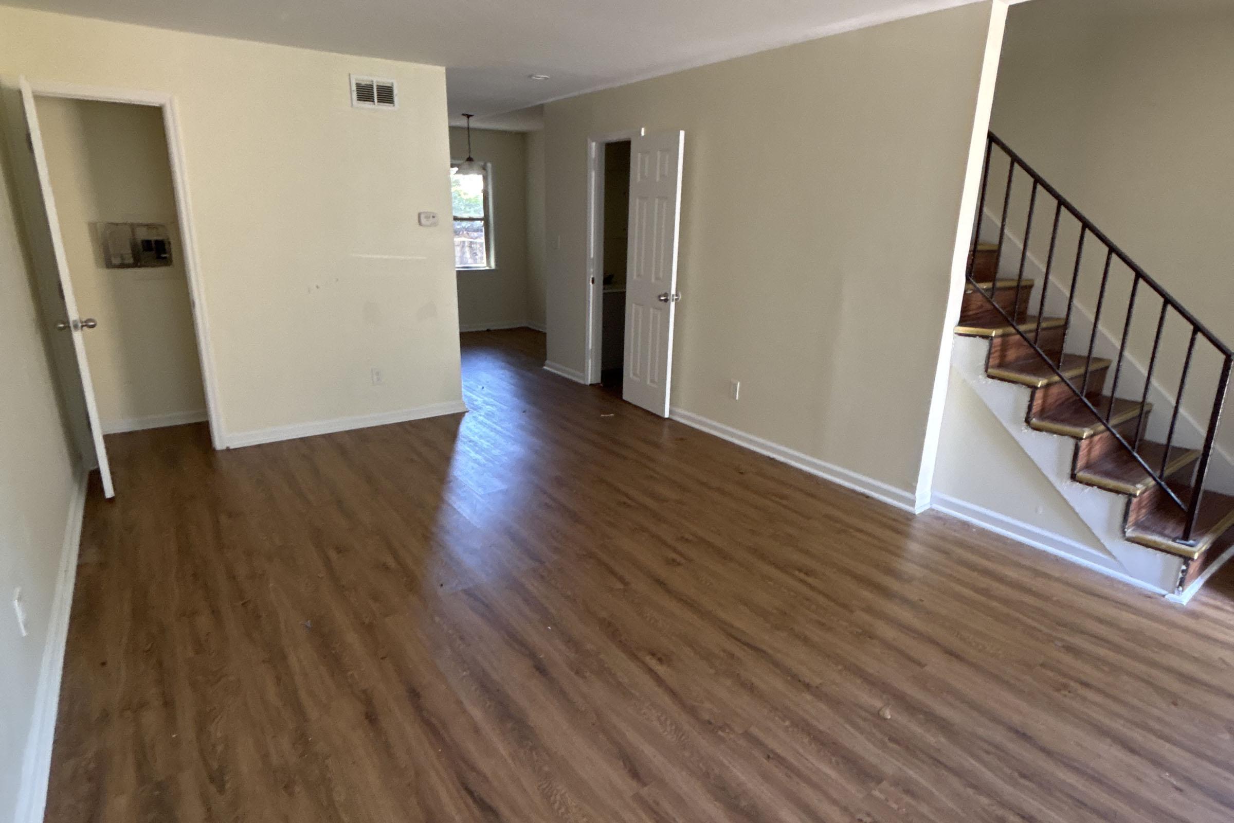 A spacious interior of a room featuring light-colored walls, hardwood flooring, and an open layout. There is a staircase on the right leading to an upper level, and two doorways leading to other rooms. Natural light enters through windows, creating a bright atmosphere.
