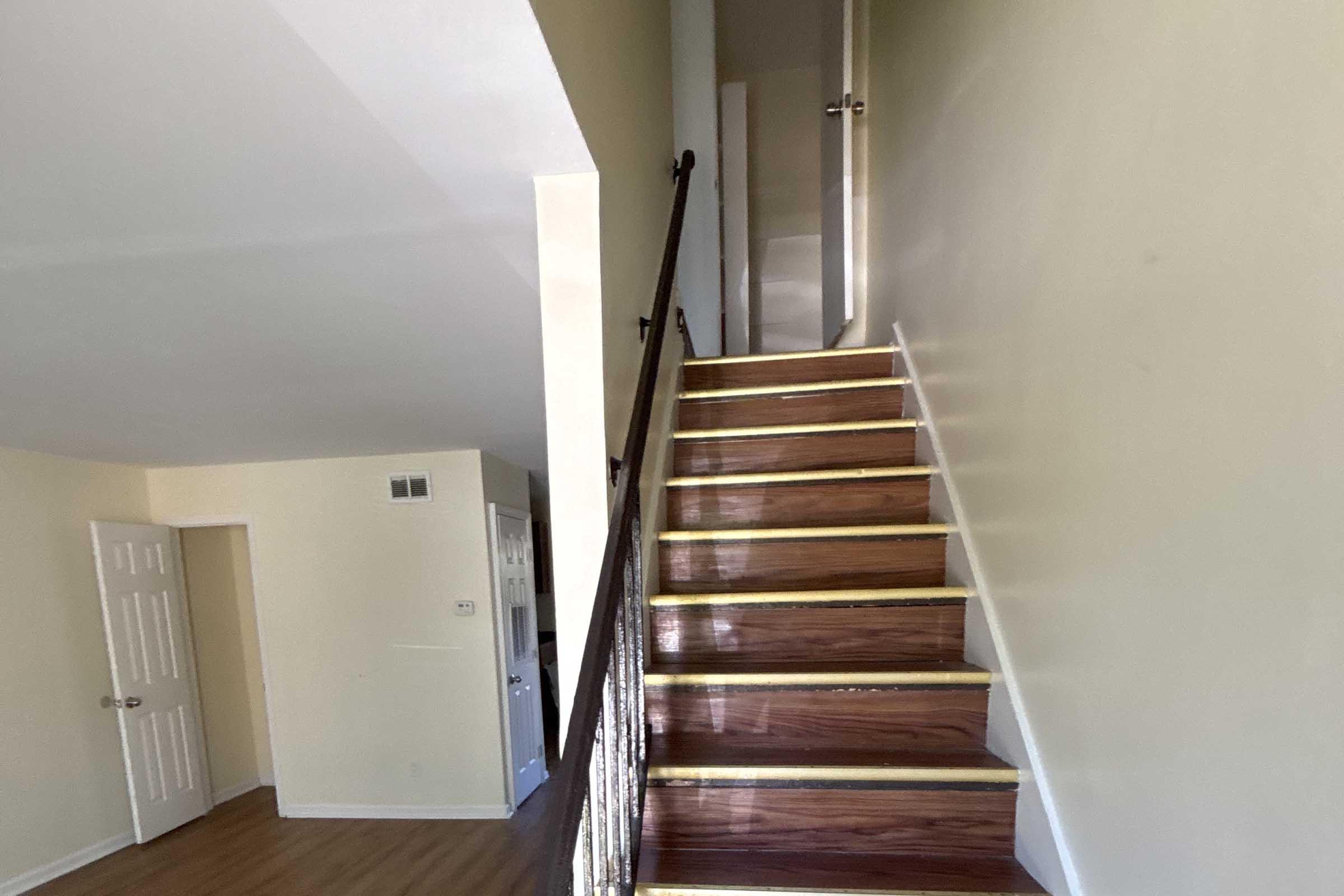 A view of an interior staircase with wooden steps and a railing, leading to an upper floor. The walls are painted light yellow, and there is a closed door at the top of the stairs. The flooring at the bottom is a light wood. A door frame is visible on the left side of the image.